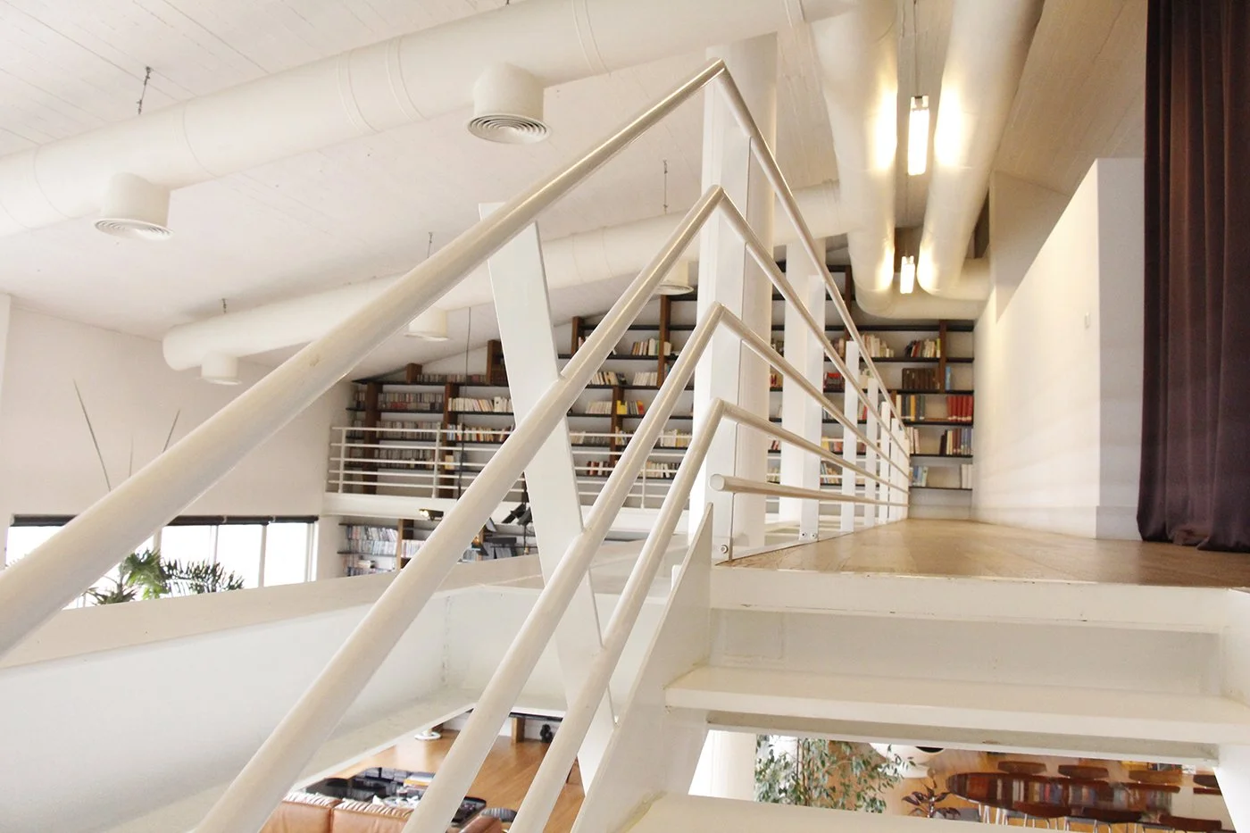 Interior view of a modern staircase with white railings leading to a second-floor library with bookshelves filled with books, beige walls, and a ceiling with exposed white ducts and lighting fixtures.
