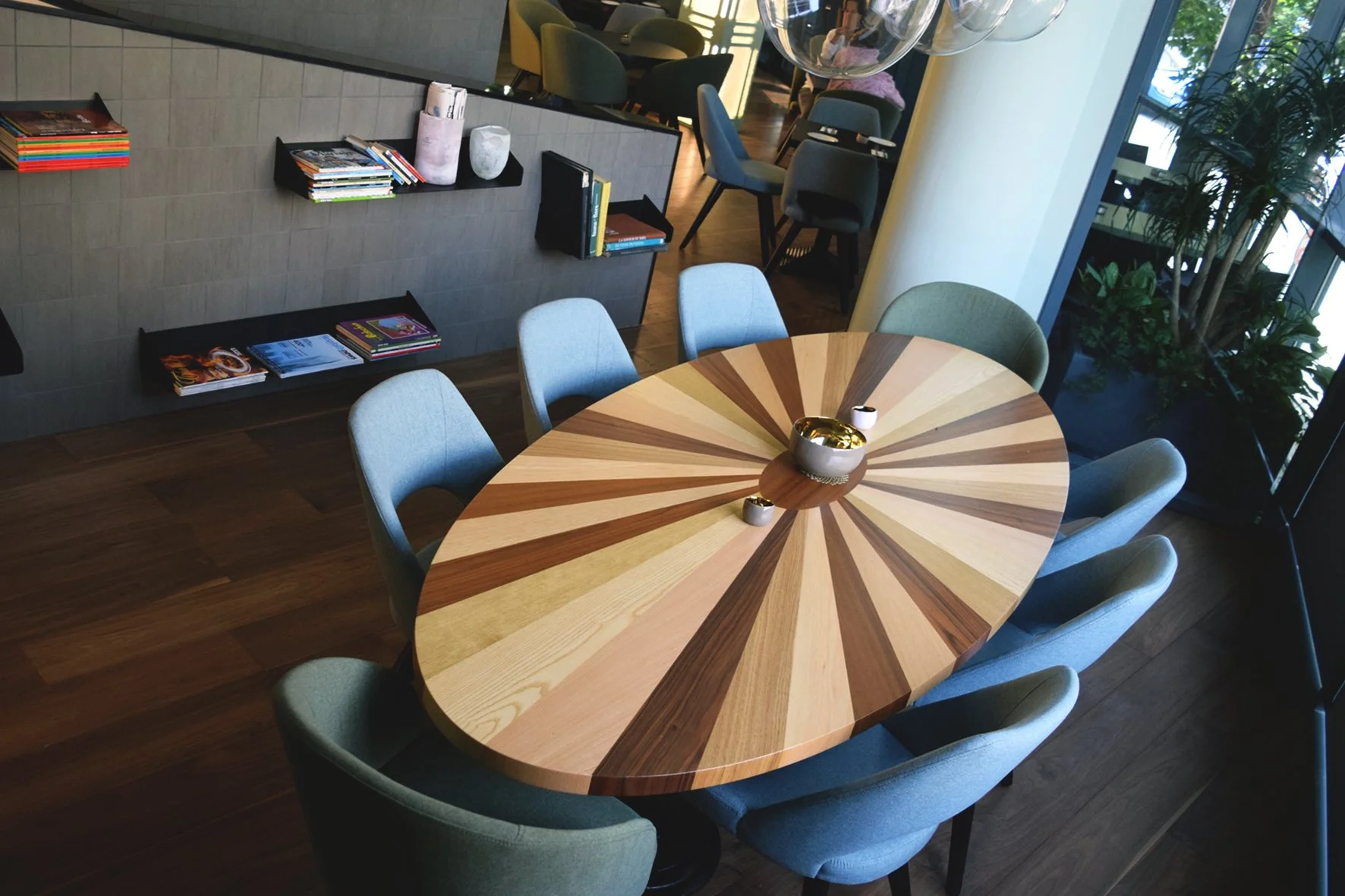 Interior of a modern restaurant or cafe with a round wooden table surrounded by eight light green chairs, with decorative vases and bowls on the table, bookshelf with magazines and books on the wall, large window with plants outside, and some dark-co