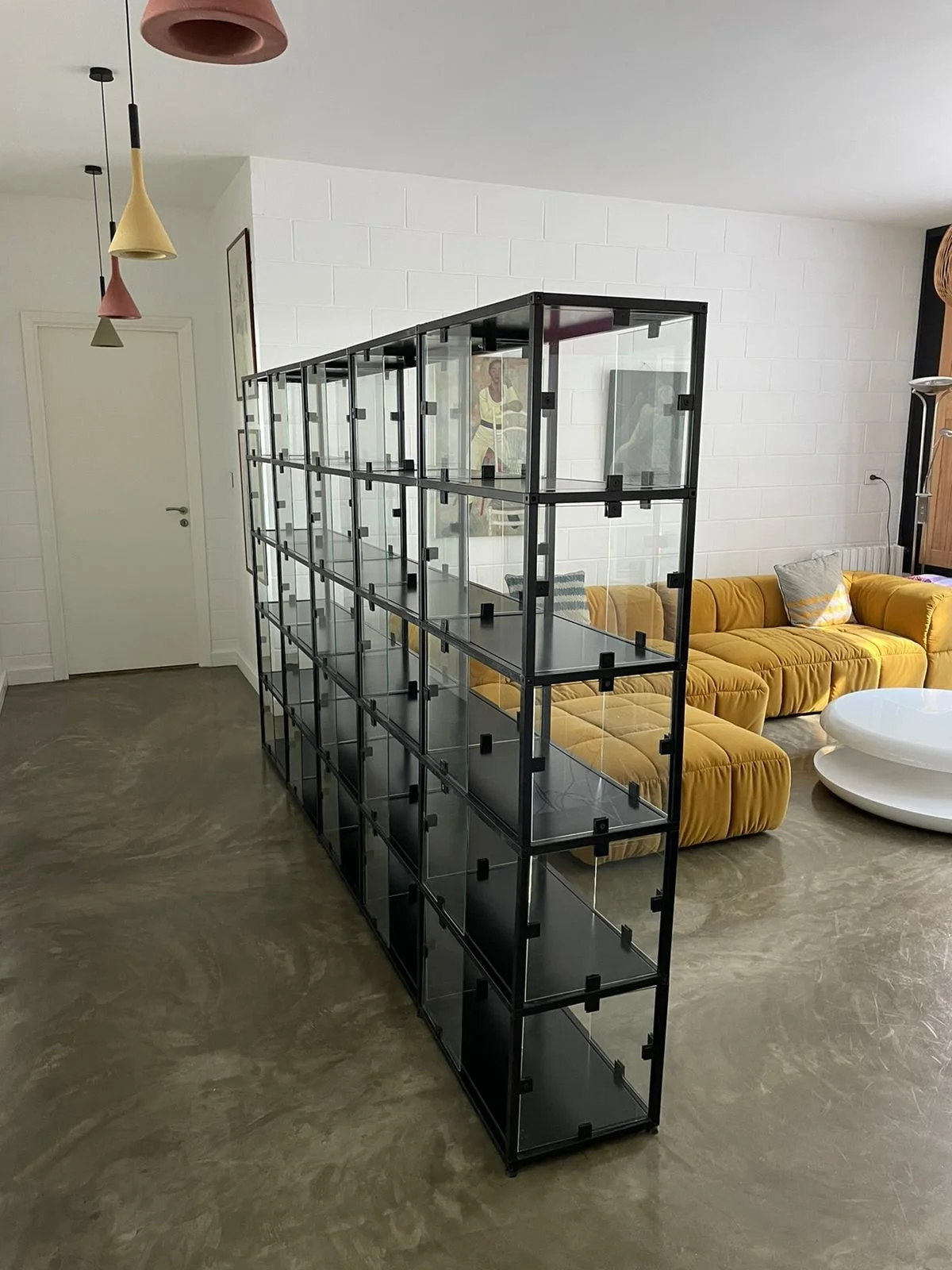 Empty black metal and glass shelving unit in a contemporary living room with yellow sofa, white coffee table, and hanging pendant lights.