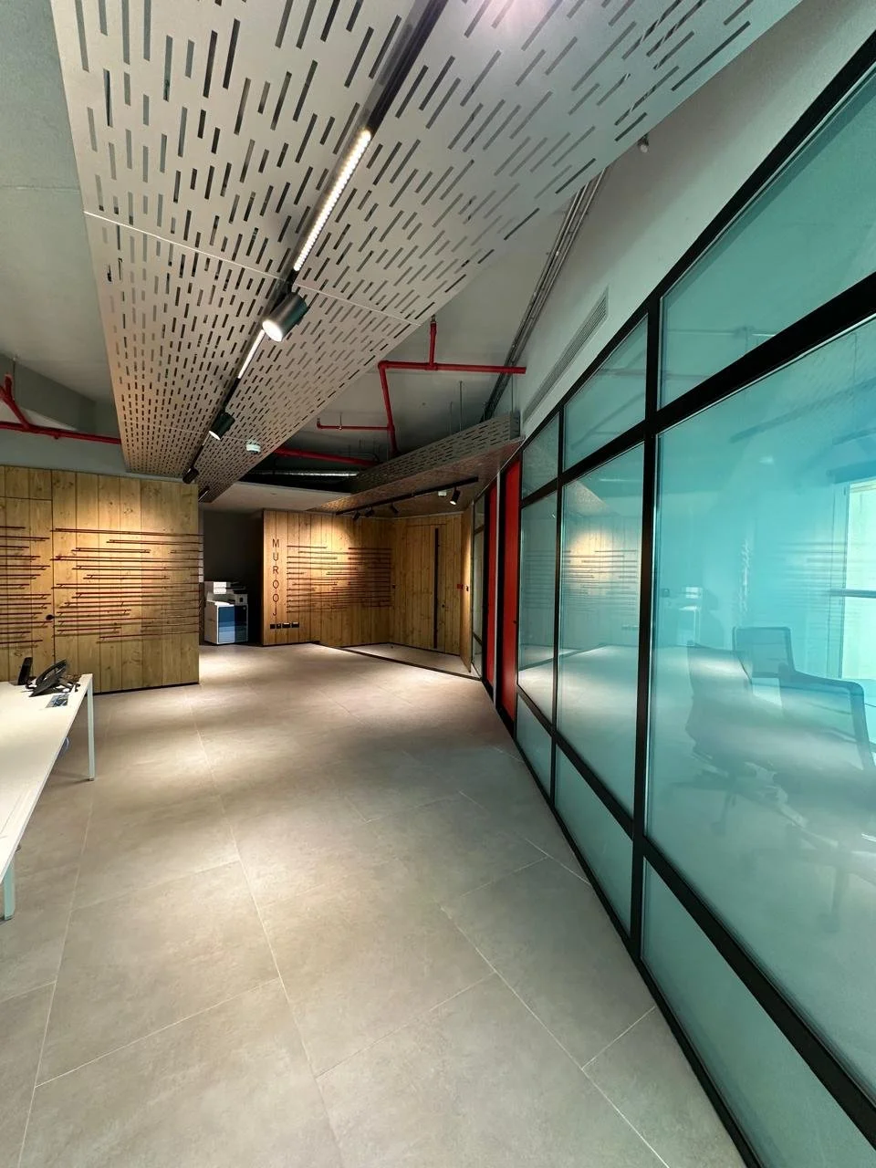 Modern office lobby with wood-paneled walls, glass partition, and ceiling panels with linear cutouts, illuminated track lighting, and red pipes on the ceiling.