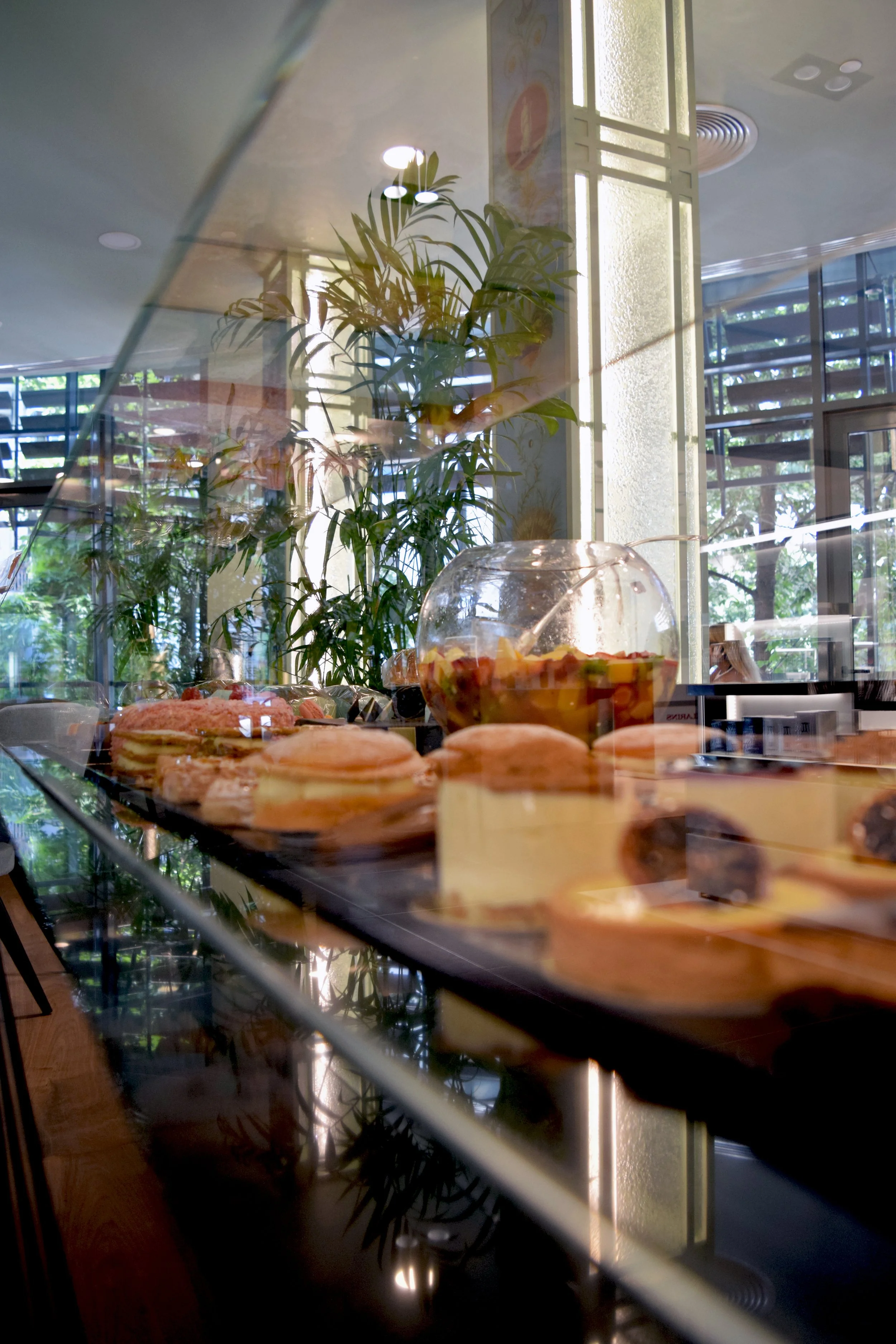 Display case with an assortment of pastries and a glass bowl of fruit salad, with a plant and large windows in the background.