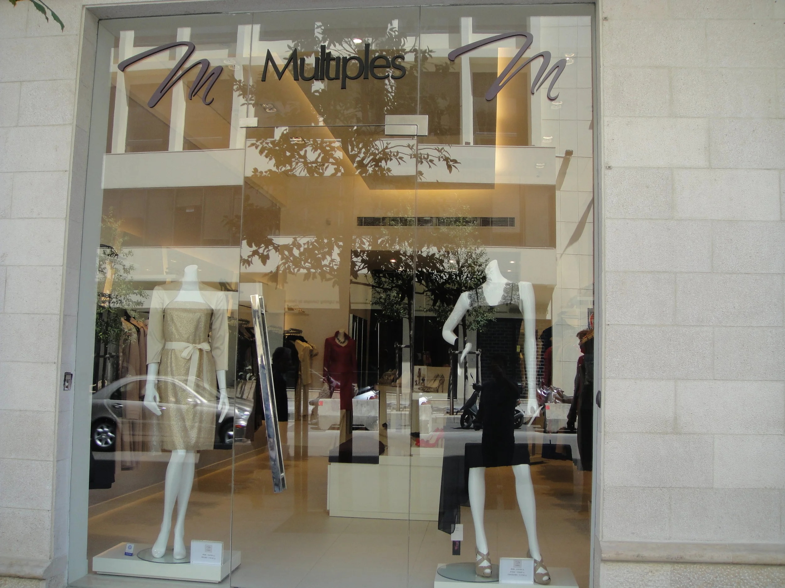 Storefront window display with mannequins dressed in elegant clothes, reflecting trees and cars outside, with the store name 'Multi' partially visible in the signage above.