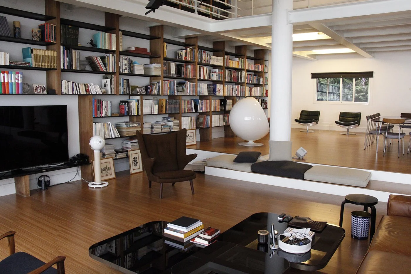 A modern living room with a large bookshelf filled with books, a flat-screen TV, a dark wooden armchair, a white egg-shaped chair, a beige cushion on the floor, and a black coffee table with books and remote controls, with a dining area and windows i