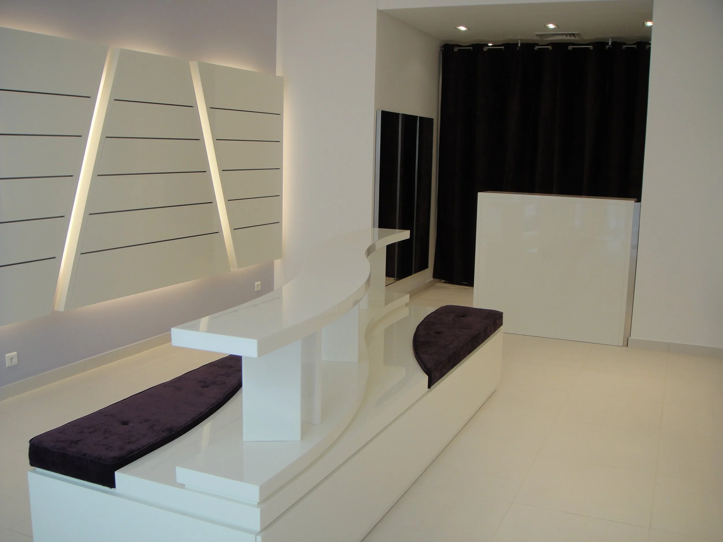 Modern white reception desk with curved edges, dark purple cushioned seating along the front, and a white shelving unit with backlit panels on the left wall. Black curtains in the background and recessed ceiling lights.