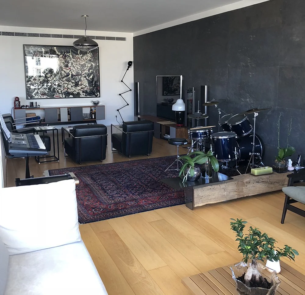 A modern living room with a black wall, contemporary furniture, an abstract artwork, a drum set on a wooden platform, potted plants, a vintage rug, and a small table with plants.