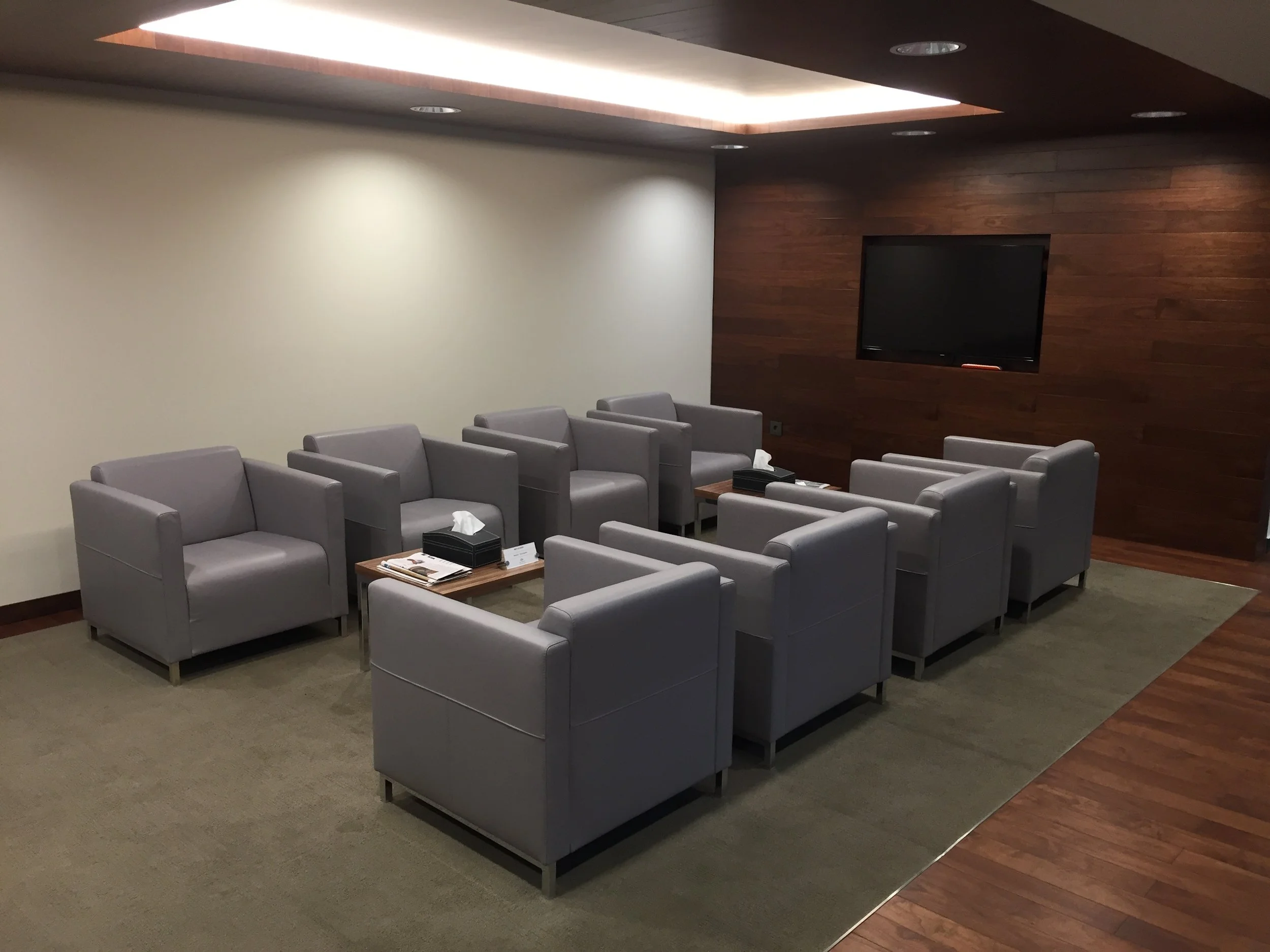 A waiting room with gray chairs arranged in two rows facing each other, a small table with tissues and papers, a large flat-screen TV mounted on a wooden-paneled wall, with neutral-colored walls and a ceiling with recessed lighting.