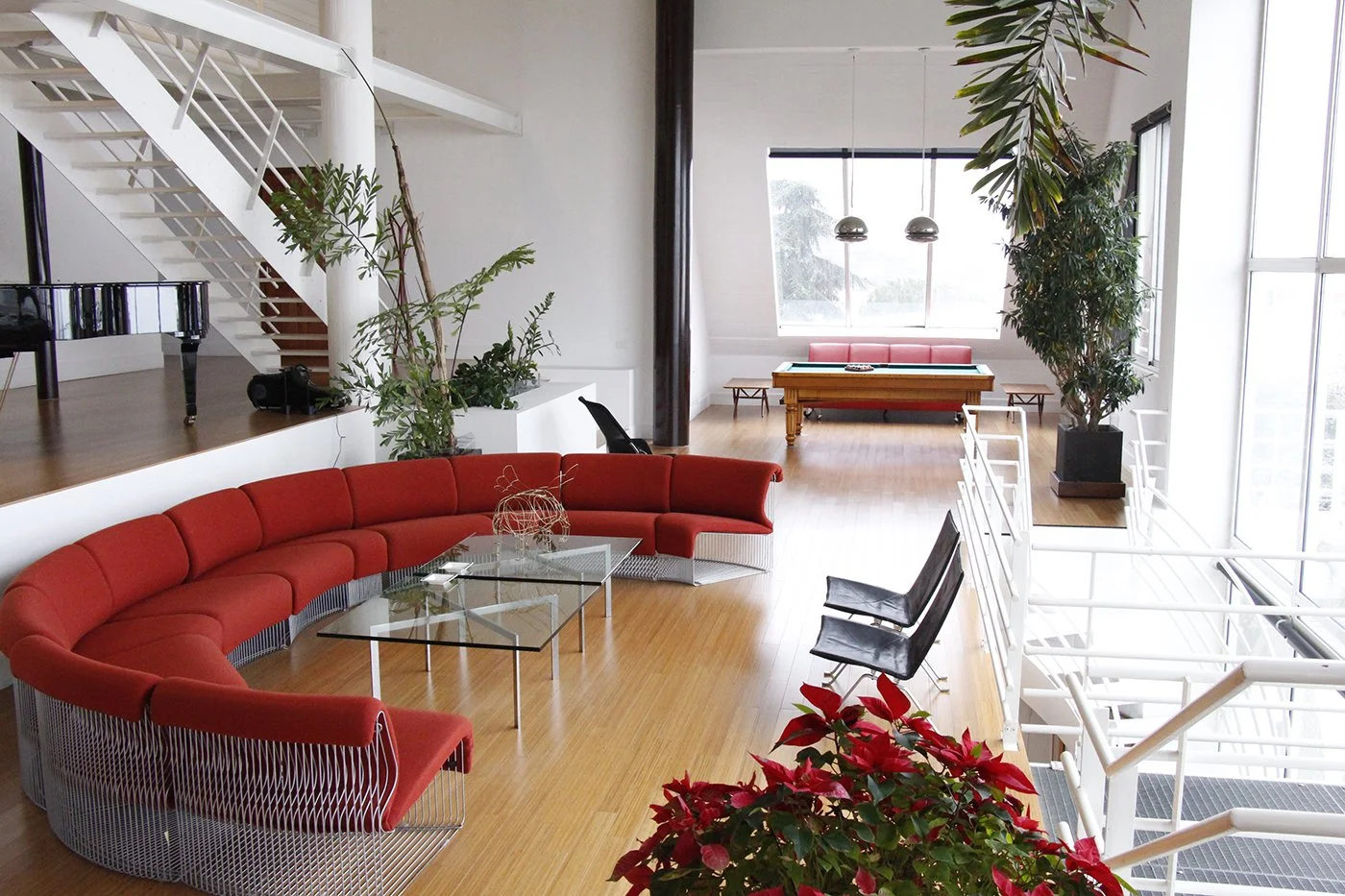 Modern living room with red curved sectional sofa, glass coffee table, and black chairs on wooden floor. Large windows and plants, staircase on left, pool table at the back, and natural light.