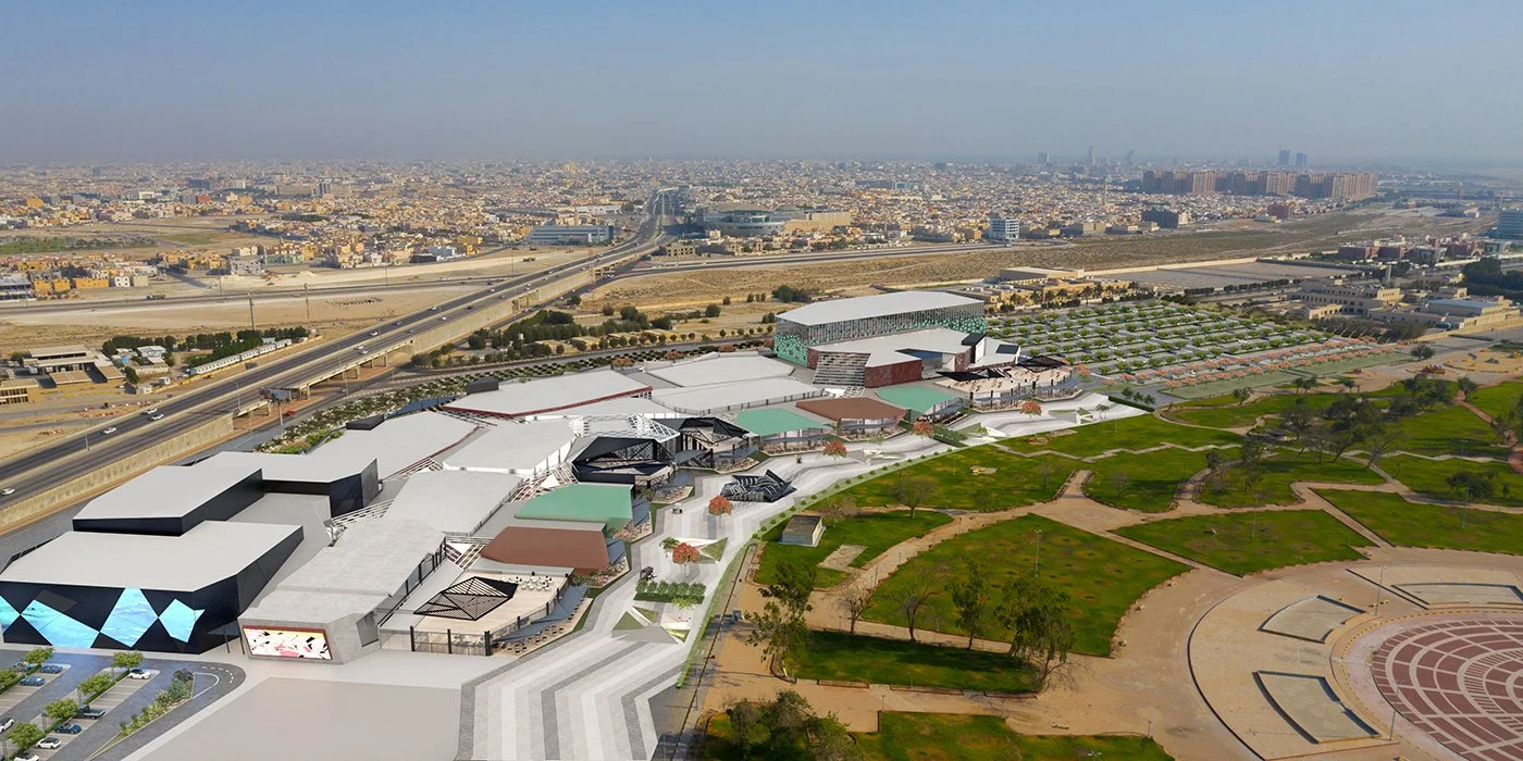 Aerial view of a modern urban complex with buildings, green spaces, and a parking lot, near highways and a densely populated city.