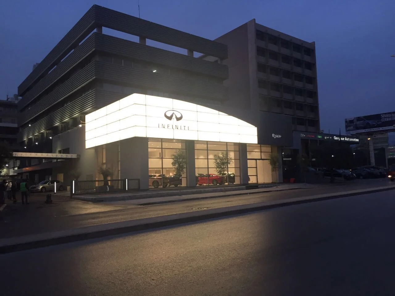 Nighttime street view of an Infiniti car dealership with illuminated signage, showing several cars inside through large glass windows, and a modern building with dark upper floors.
