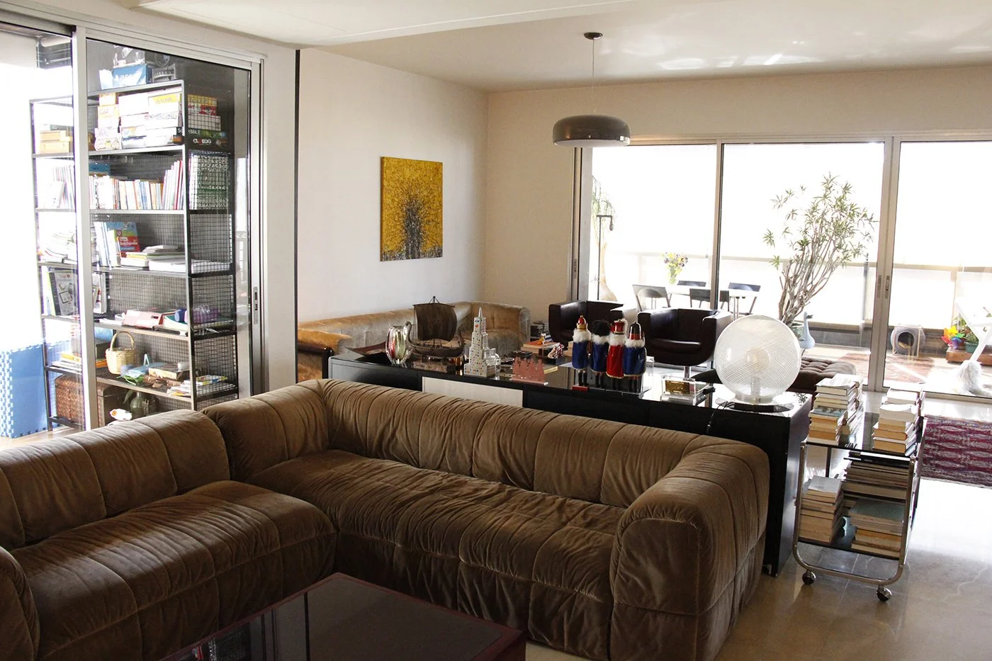 Living room with a brown sofa, black coffee table, and large glass sliding doors leading to a balcony with outdoor furniture. Decorations include books, a globe, and nutcracker figurines.