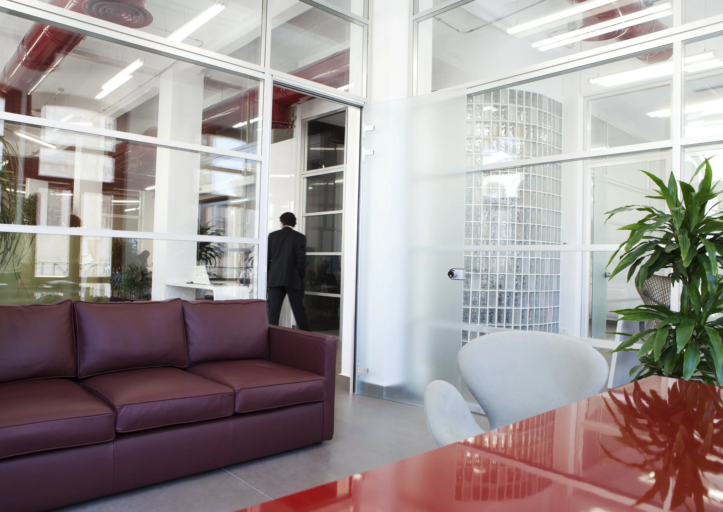Office lounge area with a burgundy leather sofa, a white chair, a red table, and a large green plant. A man in a black suit is walking through a glass door in a modern office space.