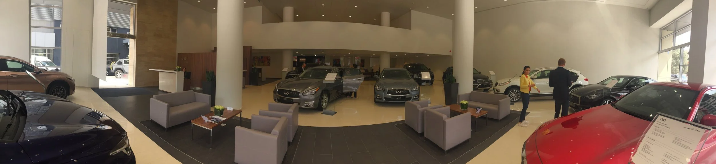 Interior of a luxury car dealership showroom with various cars on display, including a black and a red car, and a seating area with gray chairs and small tables.