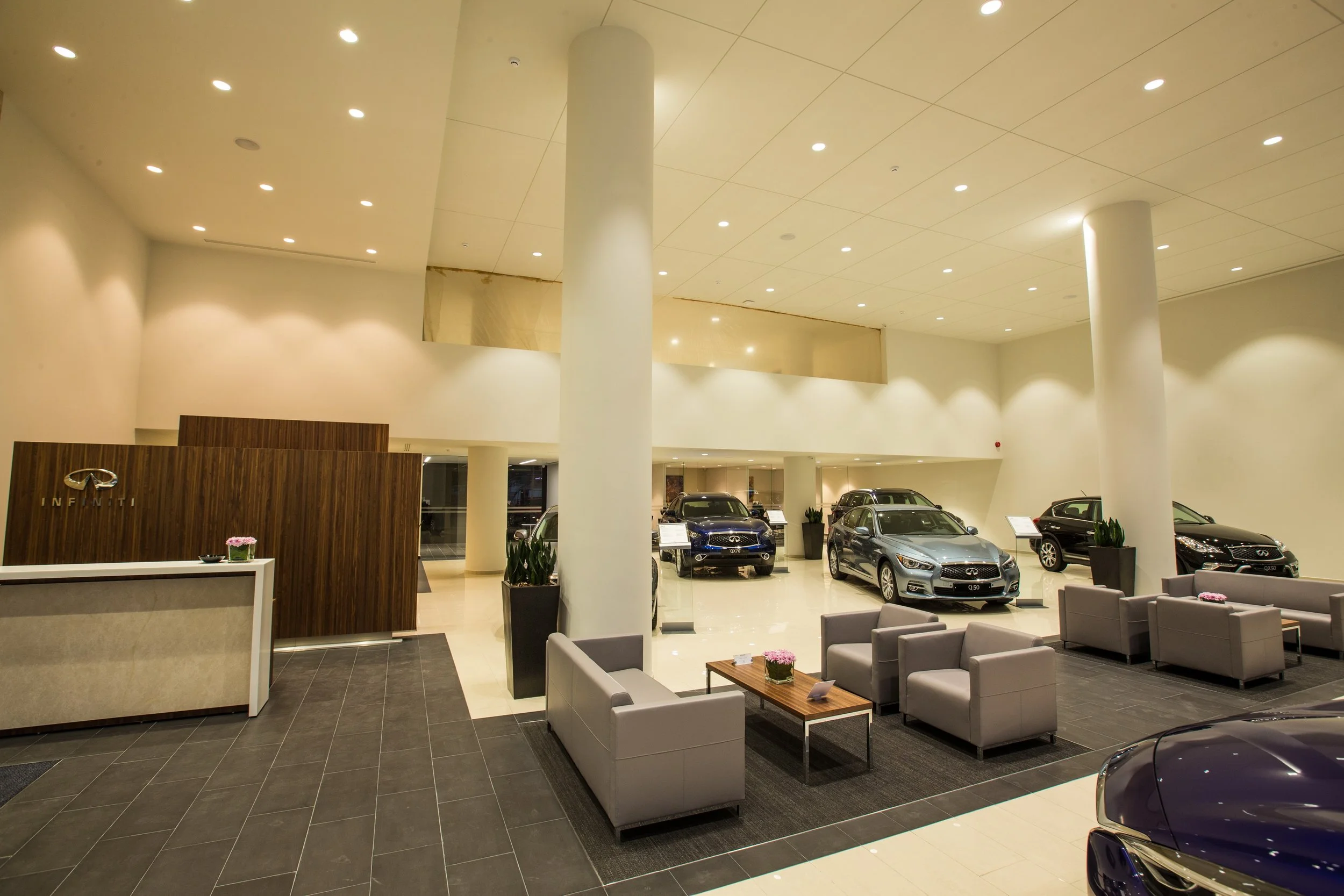 Interior view of a car dealership showroom with several modern cars on display, a reception desk, waiting area with sofas and tables, and large pillars supporting the ceiling.