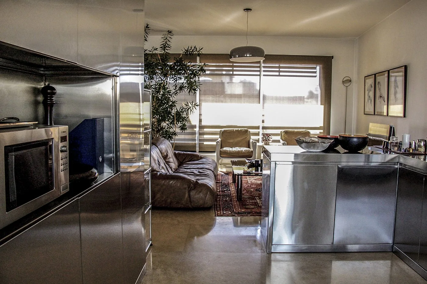 Modern loft apartment with stainless steel kitchen and living room with leather sofa and armchairs, large windows with blinds, indoor plants, and framed artwork on the wall.