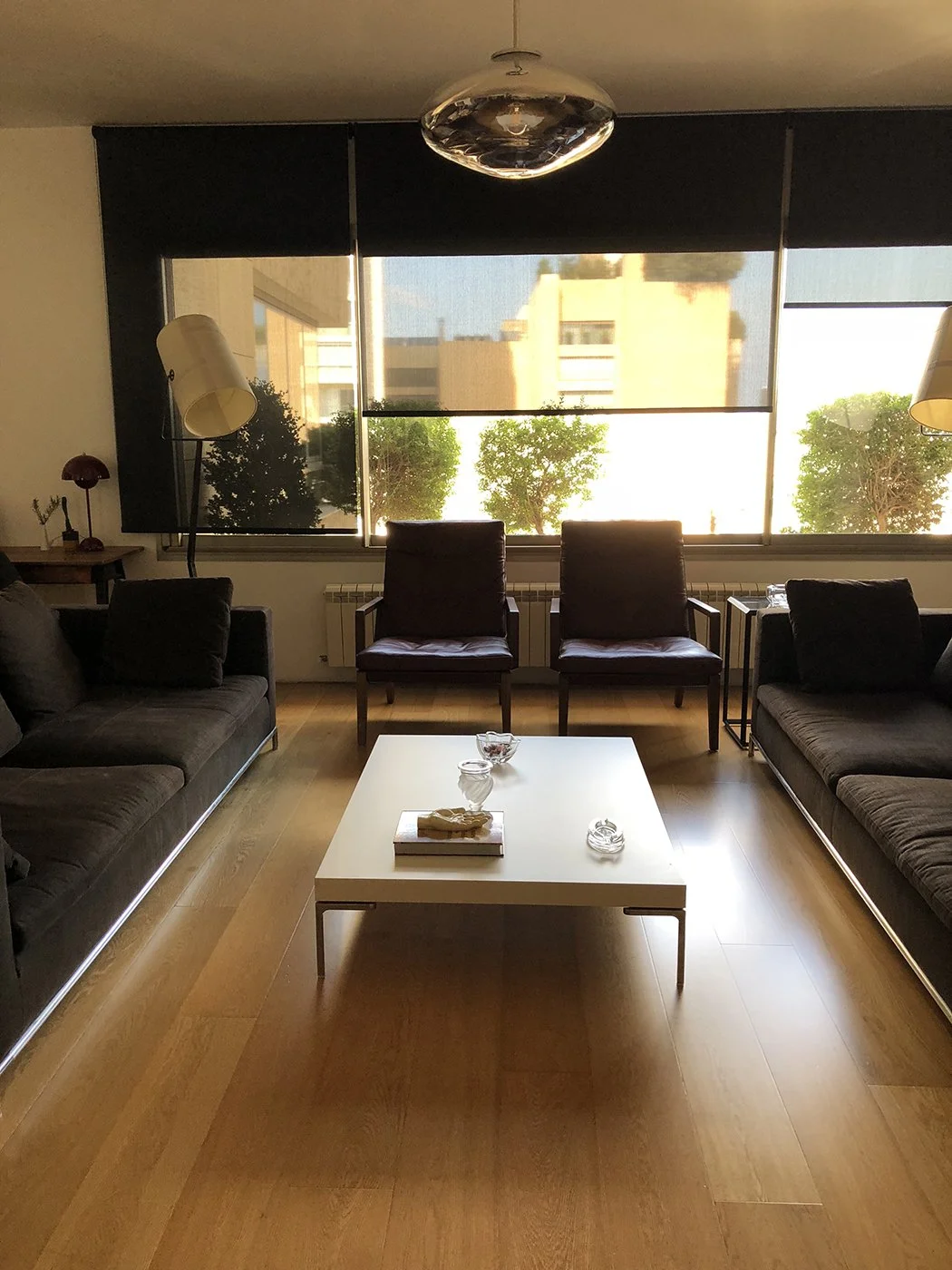 Modern living room with two dark sofas, two leather armchairs, a white coffee table with decorative items, and large windows with trees outside.