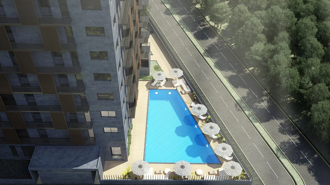 A view of a rectangular outdoor swimming pool surrounded by lounge chairs and umbrellas on a building rooftop, with a nearby road and trees in the background.