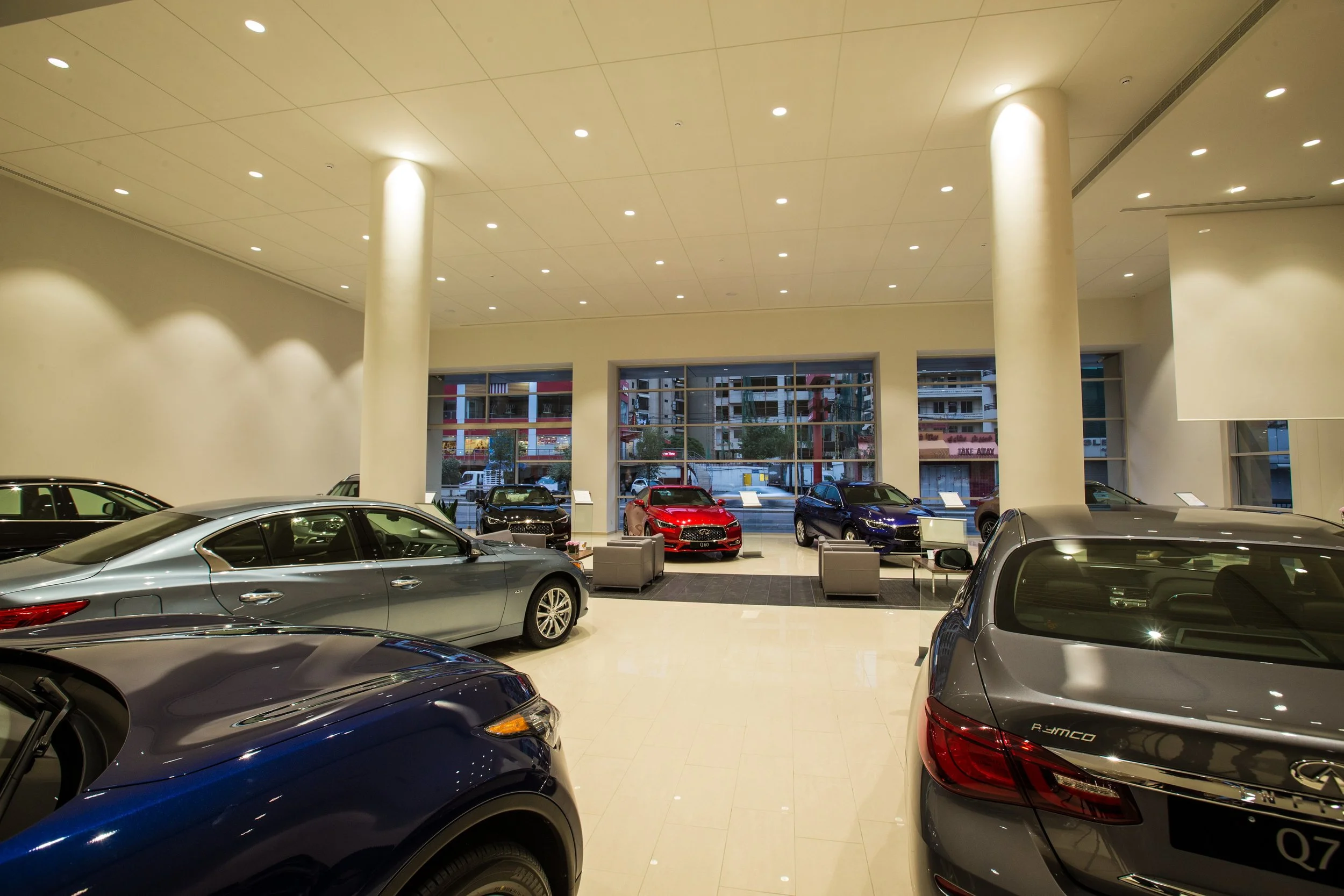 Indoor car dealership showroom with several new cars on display, large windows at the back showing buildings and street outside, bright lighting, and beige walls.
