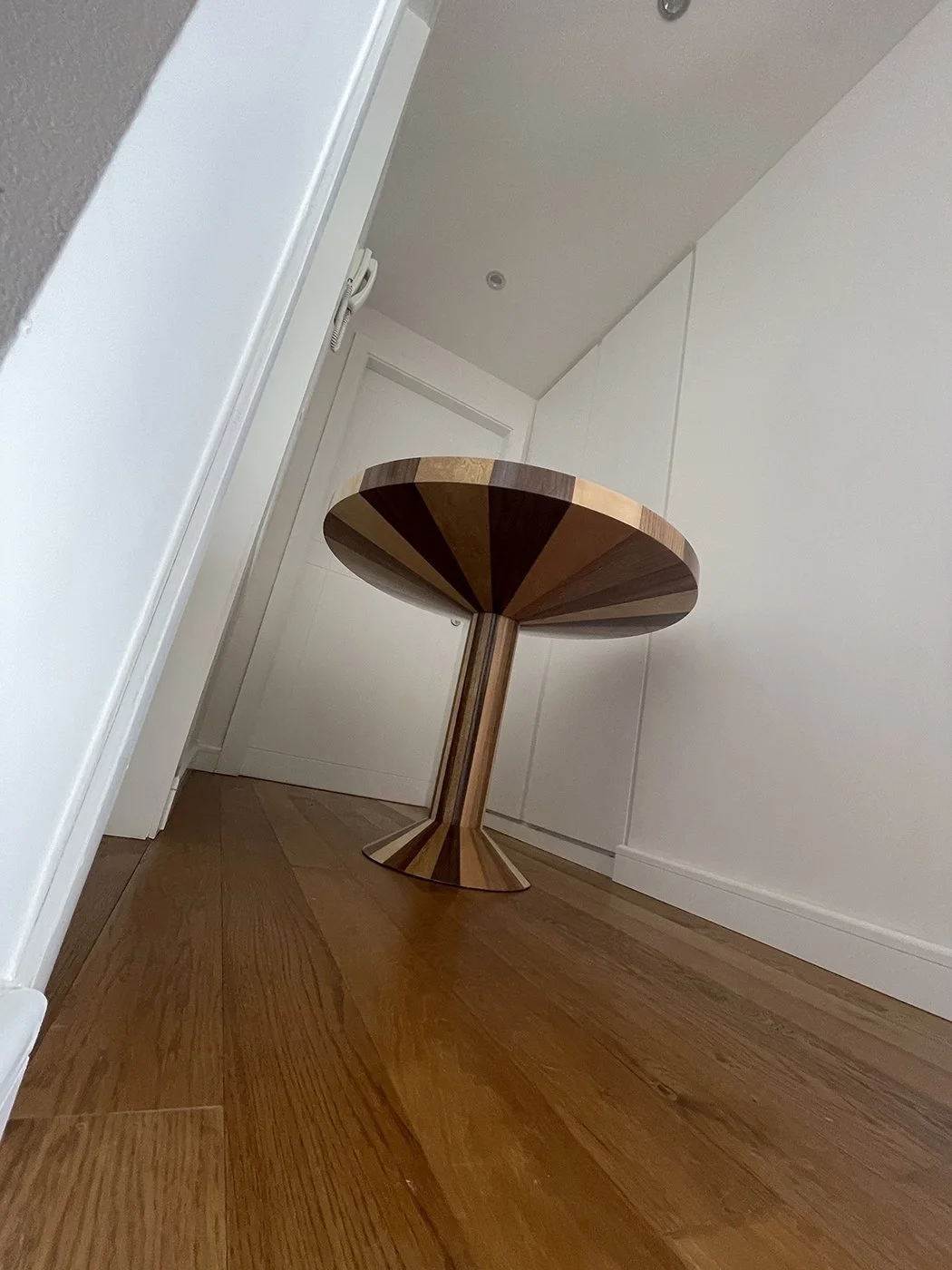 A wooden round side table with a geometric pattern on its surface, positioned in a corner of a room with hardwood flooring and white walls.