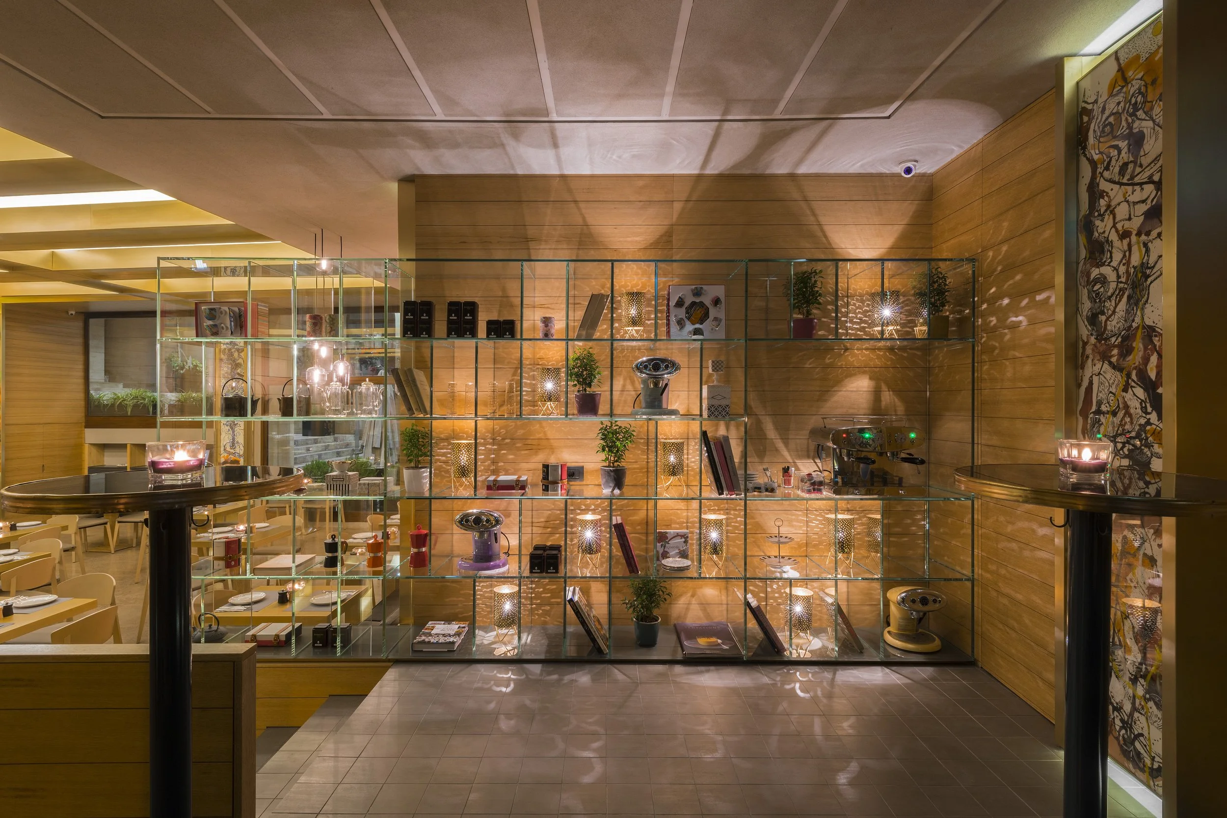 Interior view of a modern, warmly lit restaurant or cafe with a glass display shelf filled with various decorative items, plants, and books, and tables set for dining.