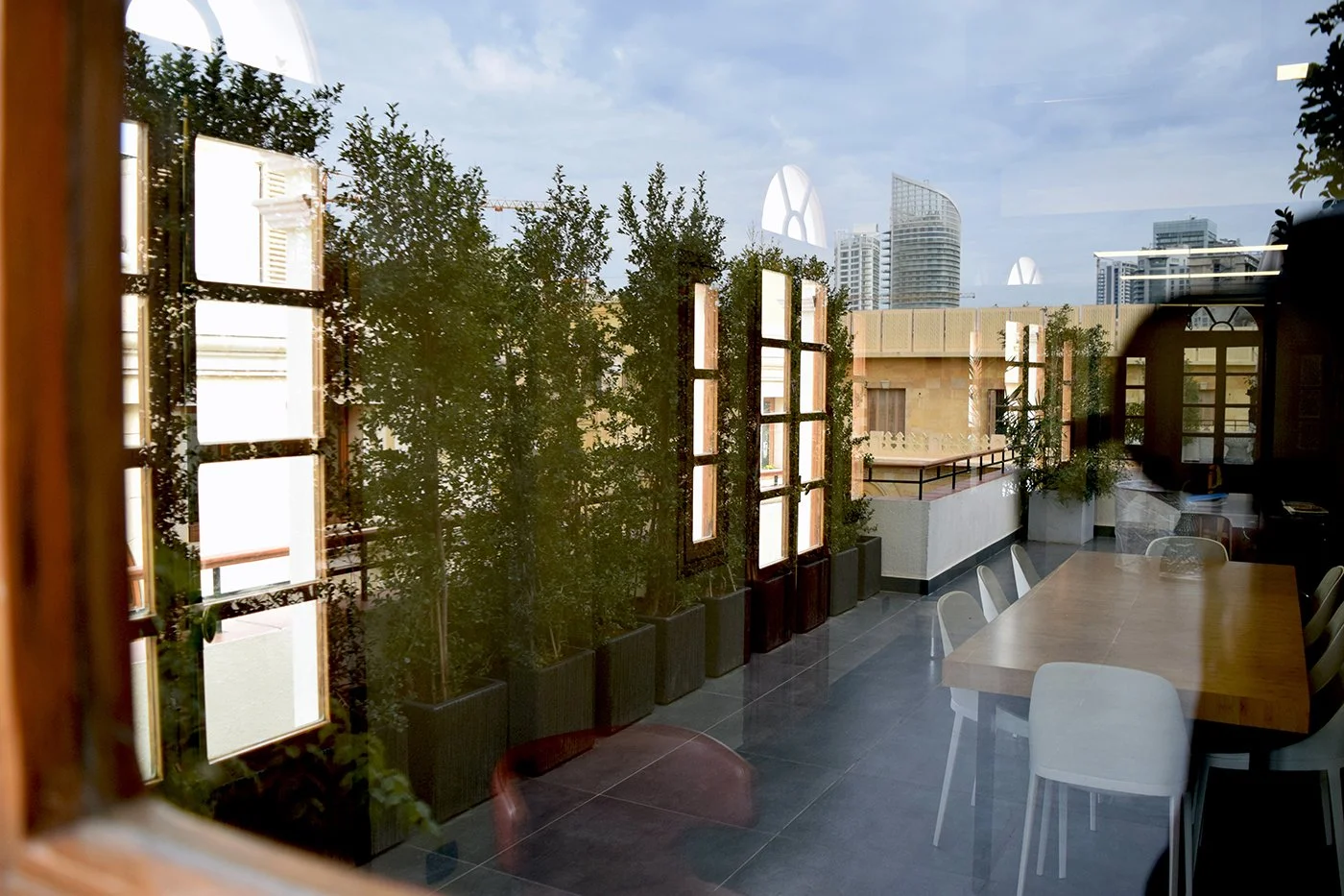 Interior view of a room with large windows, a wooden dining table, white chairs, and potted plants, with city skyline visible through the windows.