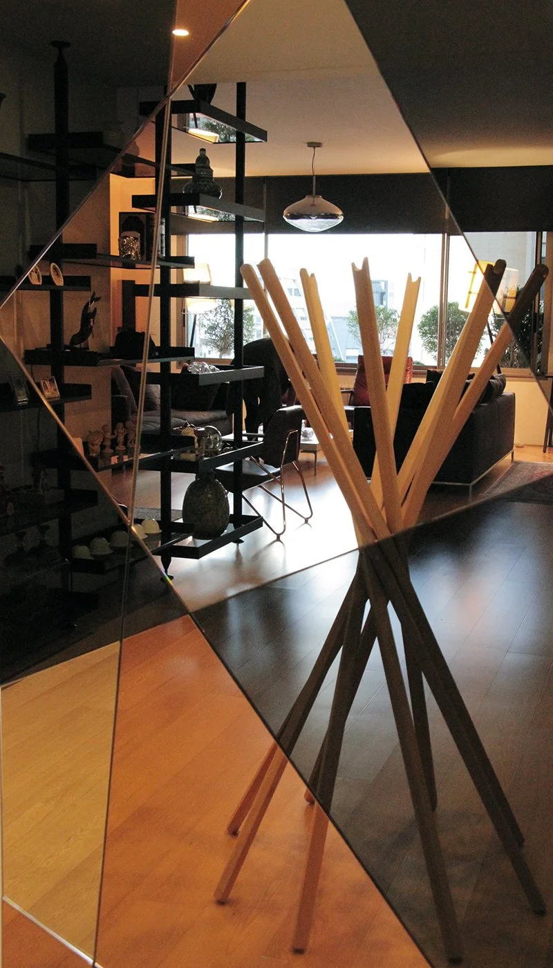 A mirror with a wooden tripod stand in a modern living room, reflecting sofas, chairs, shelves with decorative objects, and large windows showing trees and buildings outside.