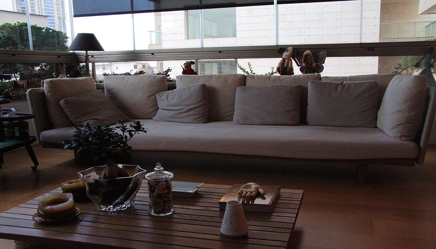 A modern living room with a large white sofa, pillows, a wooden coffee table with decorative items, plants, and plush toys on the window ledge. Large window with a city view in the background.