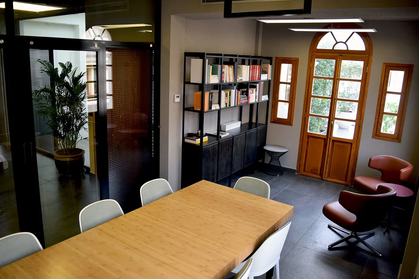 Interior of a modern office or meeting room with a wooden table, white chairs, a black bookshelf filled with books, a large window with wooden frames, and cozy chairs near the window.