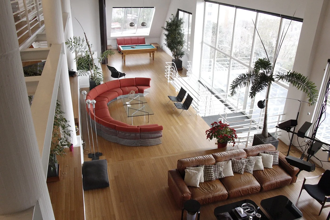 A spacious modern living room with hardwood floors, large windows, green plants, a curved red sofa, a glass coffee table, black chairs, a brown leather sofa with pillows, a pool table, and a staircase with white railings.