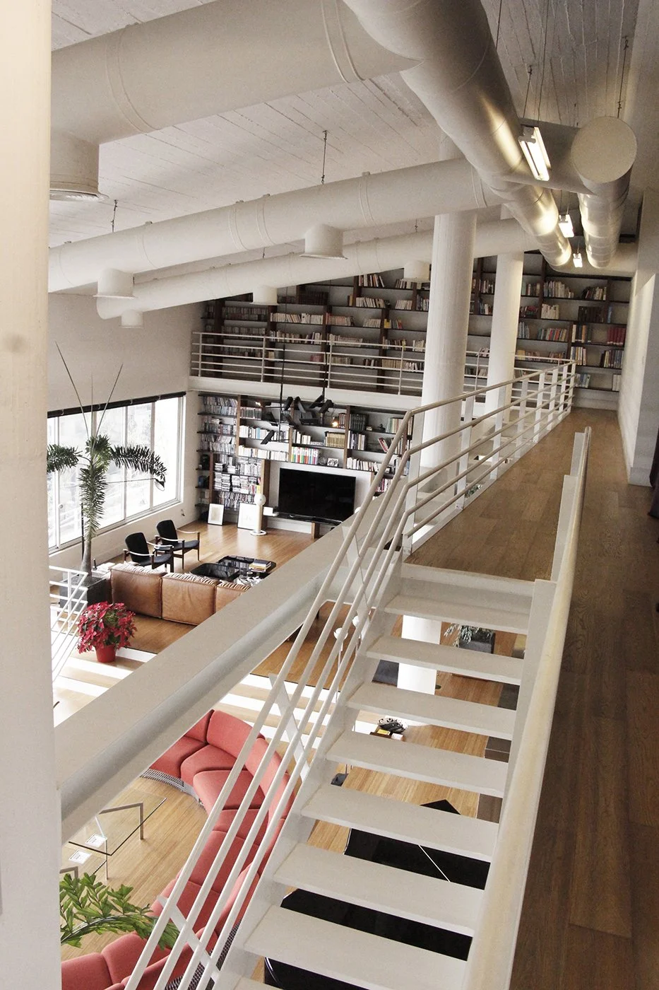 Interior of a modern, multi-level home with a living room featuring a large bookshelf, television, seating area, and decor, viewed from a staircase balcony.