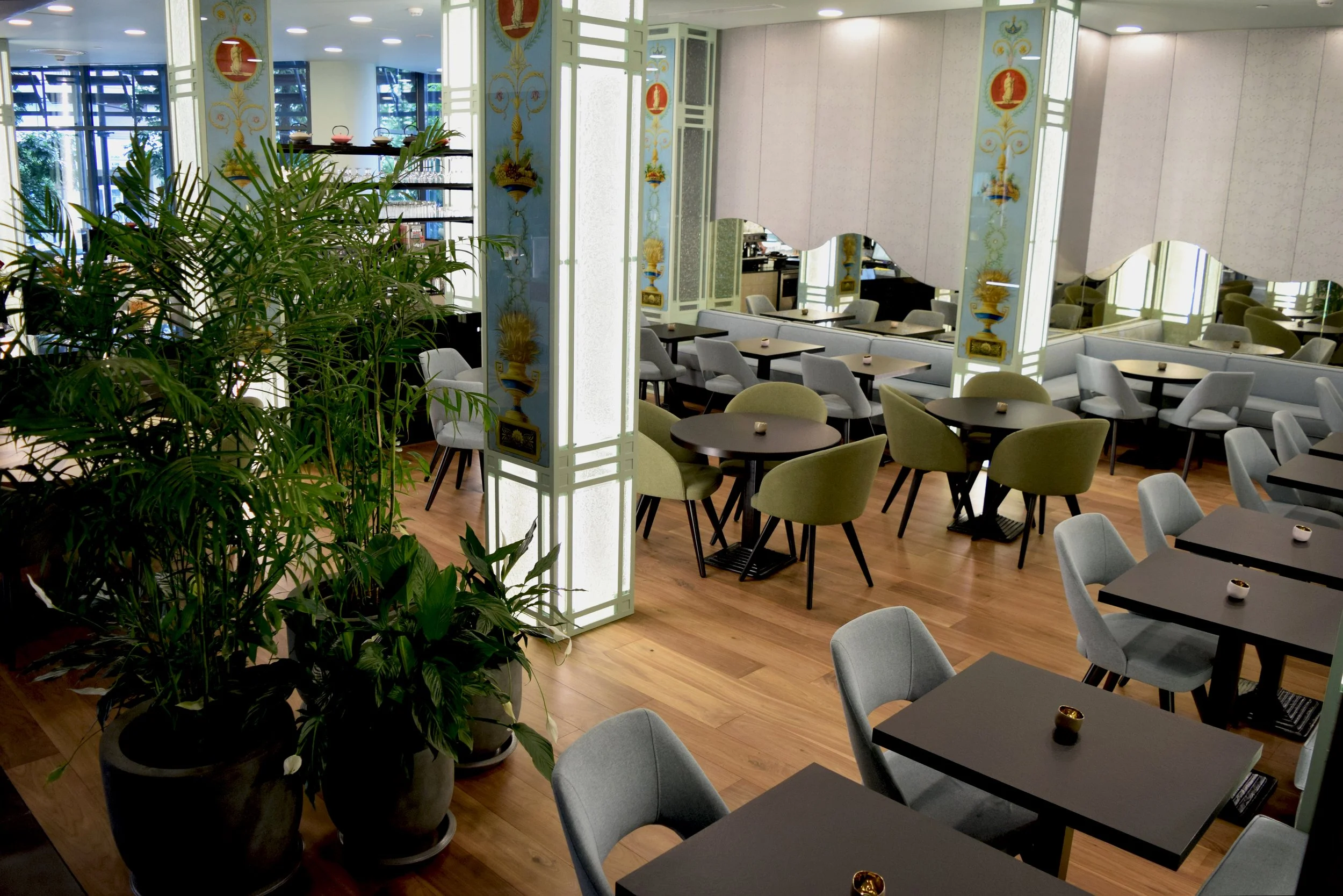 A restaurant or cafe interior with multiple tables and chairs, some with green and gray upholstery, and decorative panels with gold and blue patterns. A large green potted plant is on the left side of the image, partially obscuring the view.