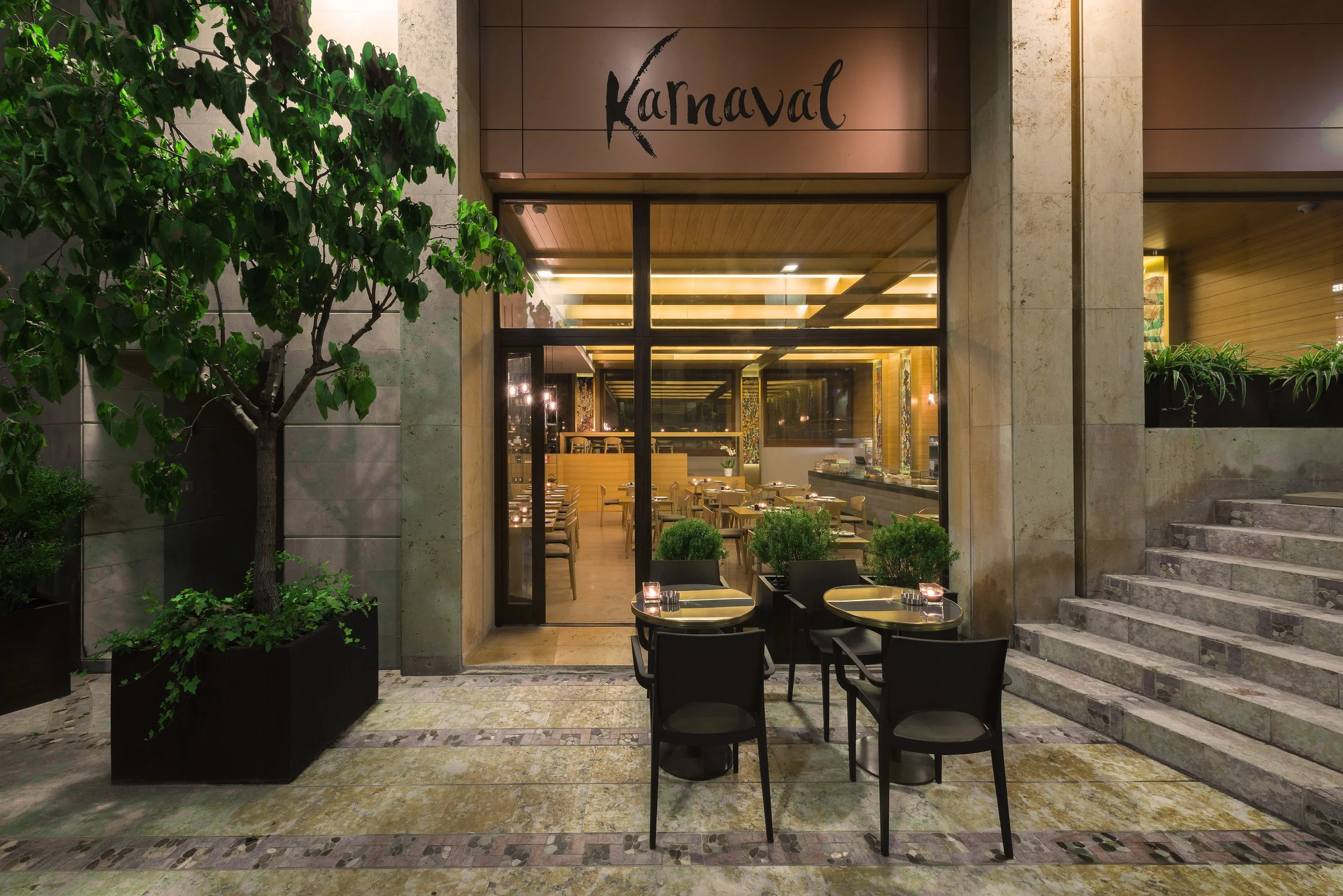 Outside view of a restaurant named 'Karnaval' with large windows revealing a warmly lit interior, surrounded by potted plants and trees, with outdoor seating on a stone patio.