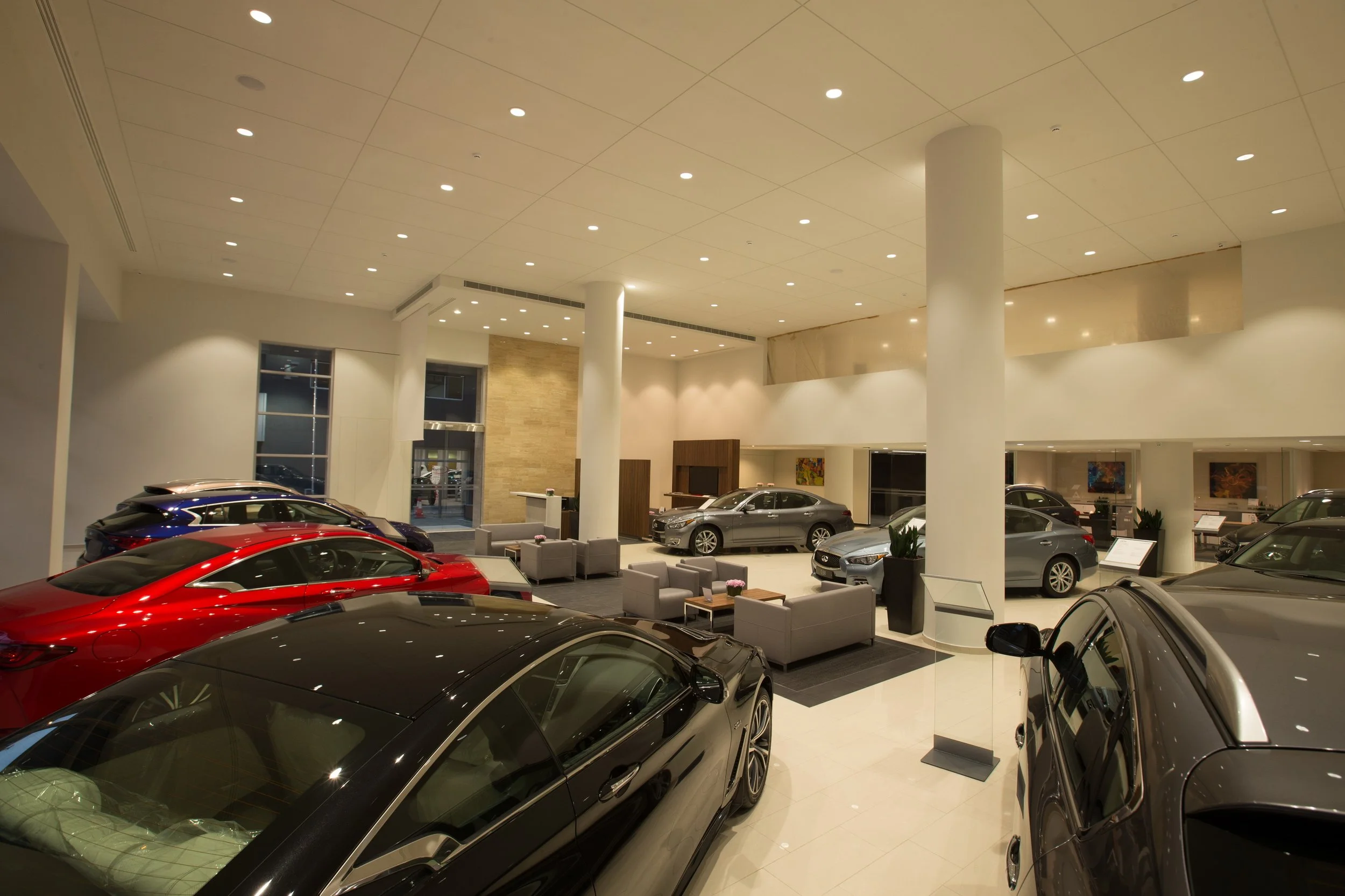 A modern car dealership showroom with multiple luxury vehicles on display, seating area, and large windows.