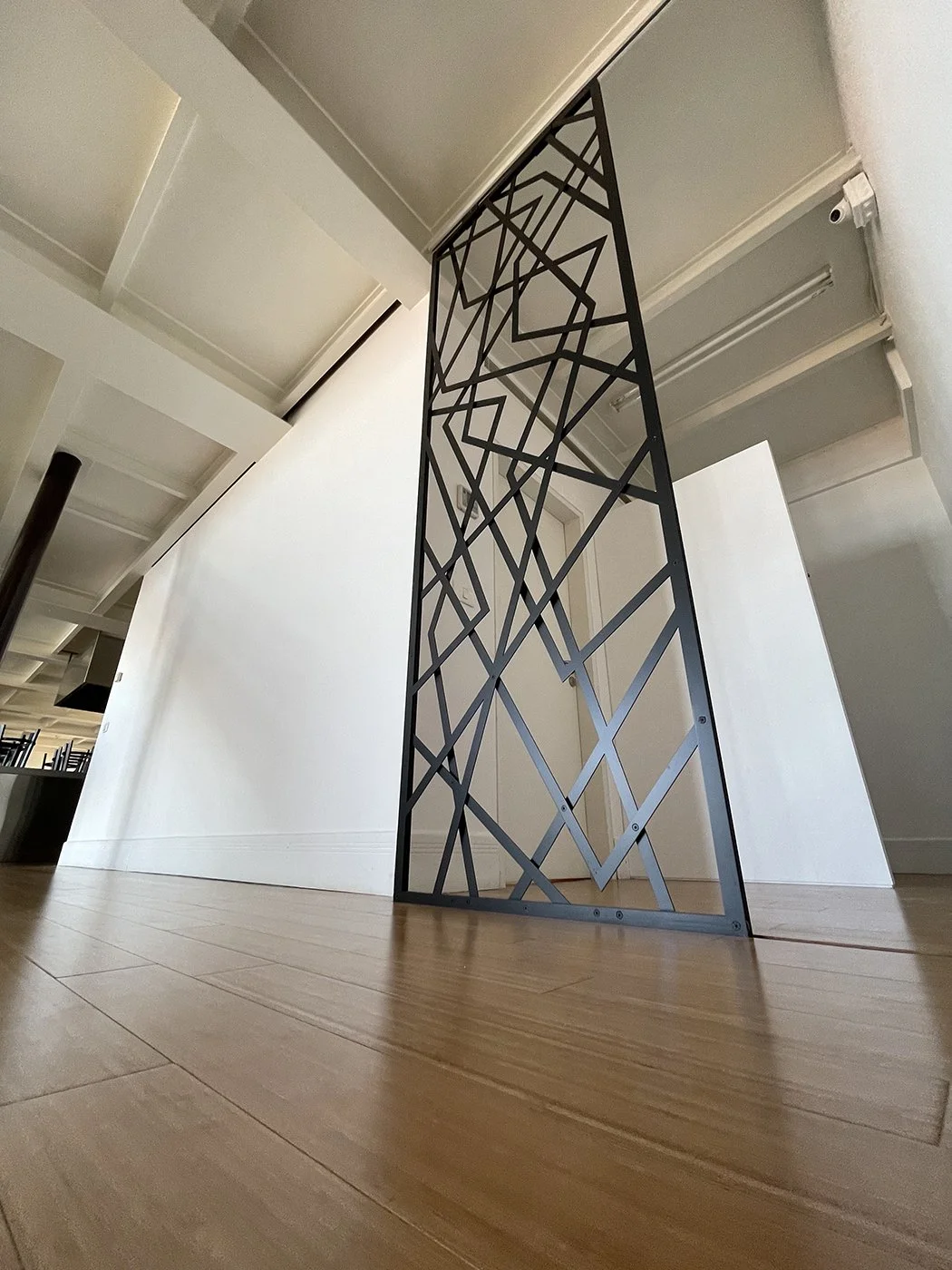 A black metal decorative room divider with geometric patterns, standing in a modern interior space with light wood flooring and white walls.