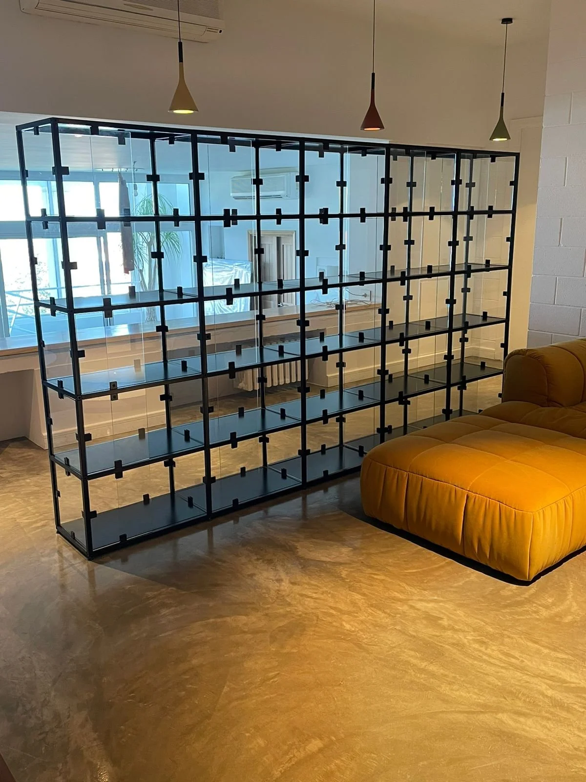 A black metal and glass bookshelf dividing a living room, with a mustard yellow sofa nearby, in front of a large window with cityscape view.