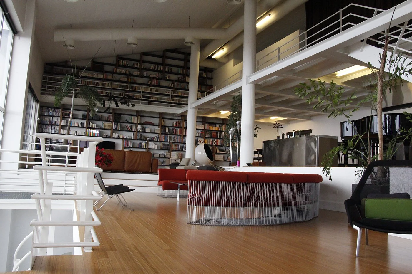 Interior of a modern building with a large bookshelf, lounge chairs, plants, and a spiral staircase.