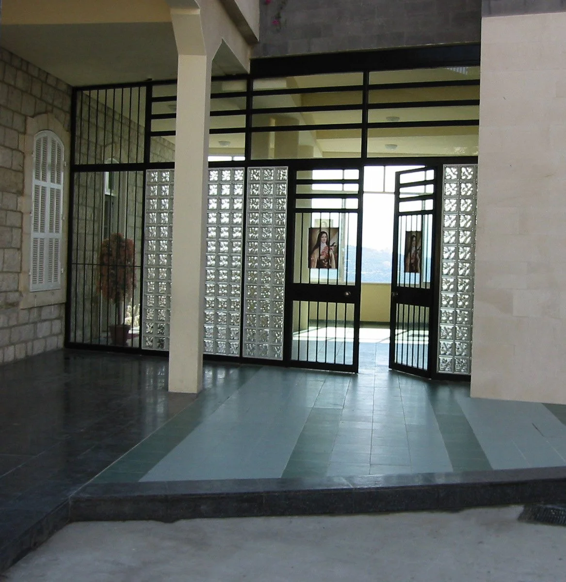 Entrance area with black metal gate and glass blocks, stone wall, potted plant, and portraits of women on the walls in the background.