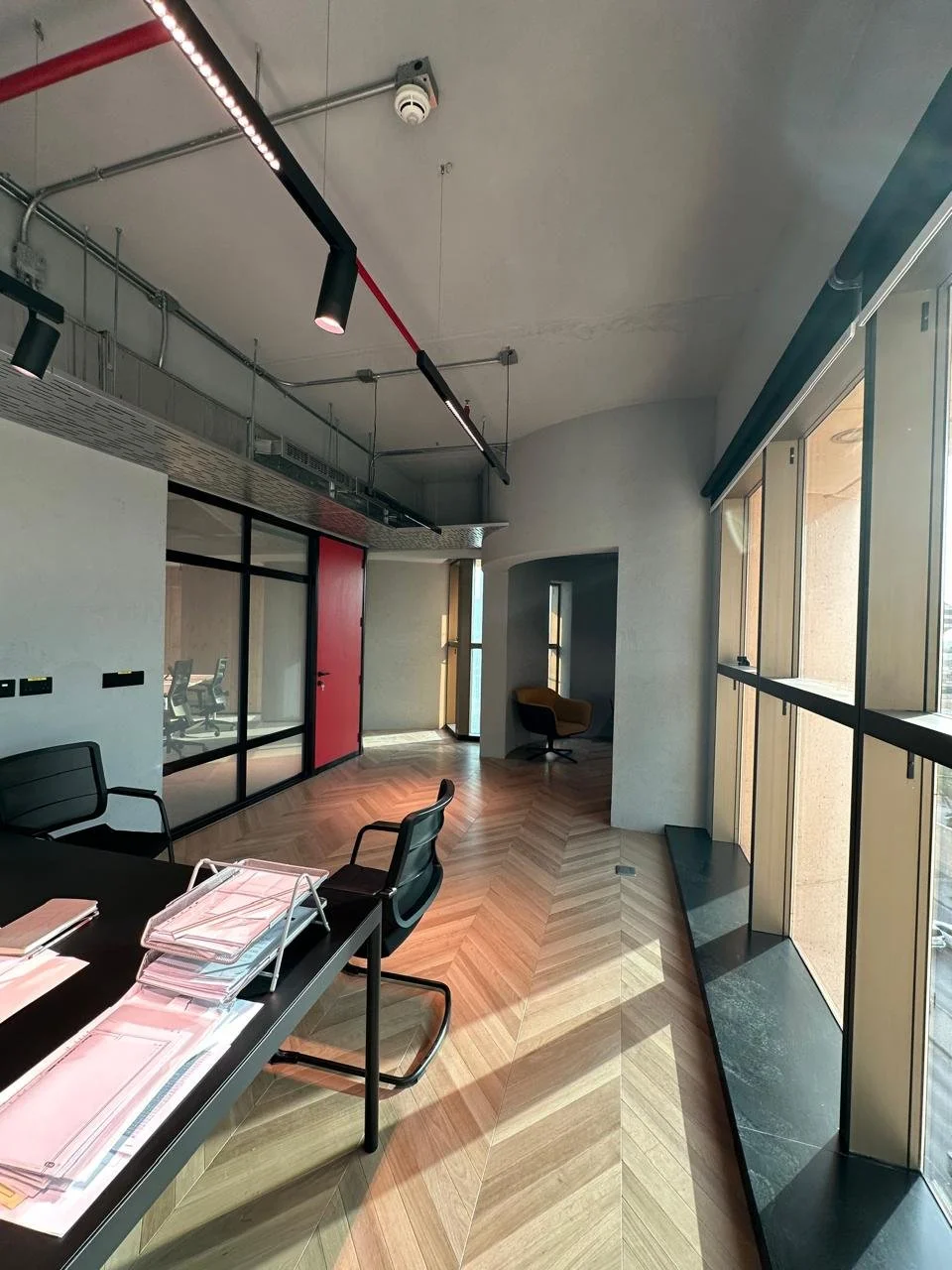 Modern conference room with black table, chairs, and large windows providing natural light, wooden floor, and a red door.