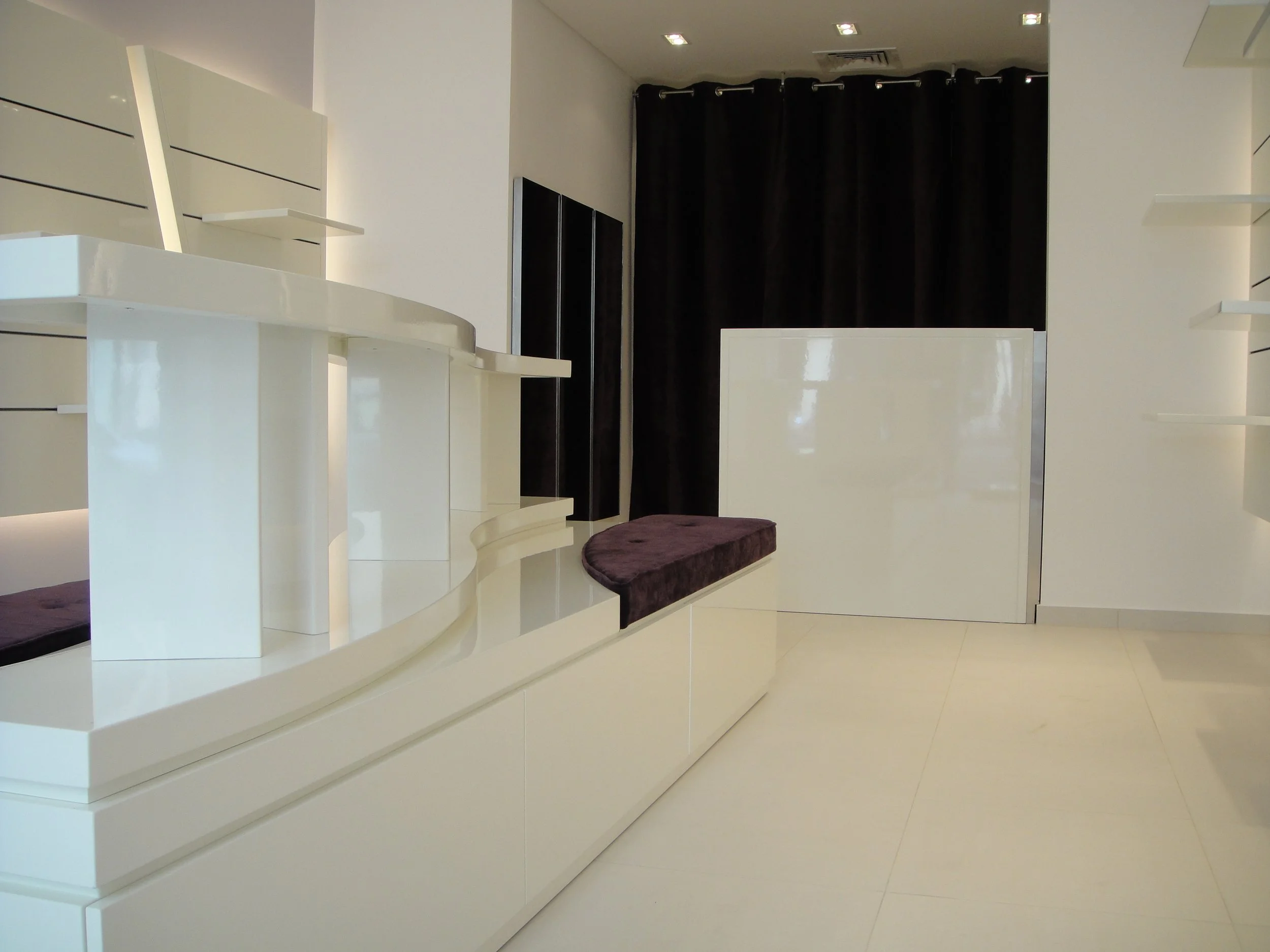 Modern white reception desk or display counter with built-in shelving and seating area, in a bright and minimalistic interior space with black curtains in the background and ceiling lights.