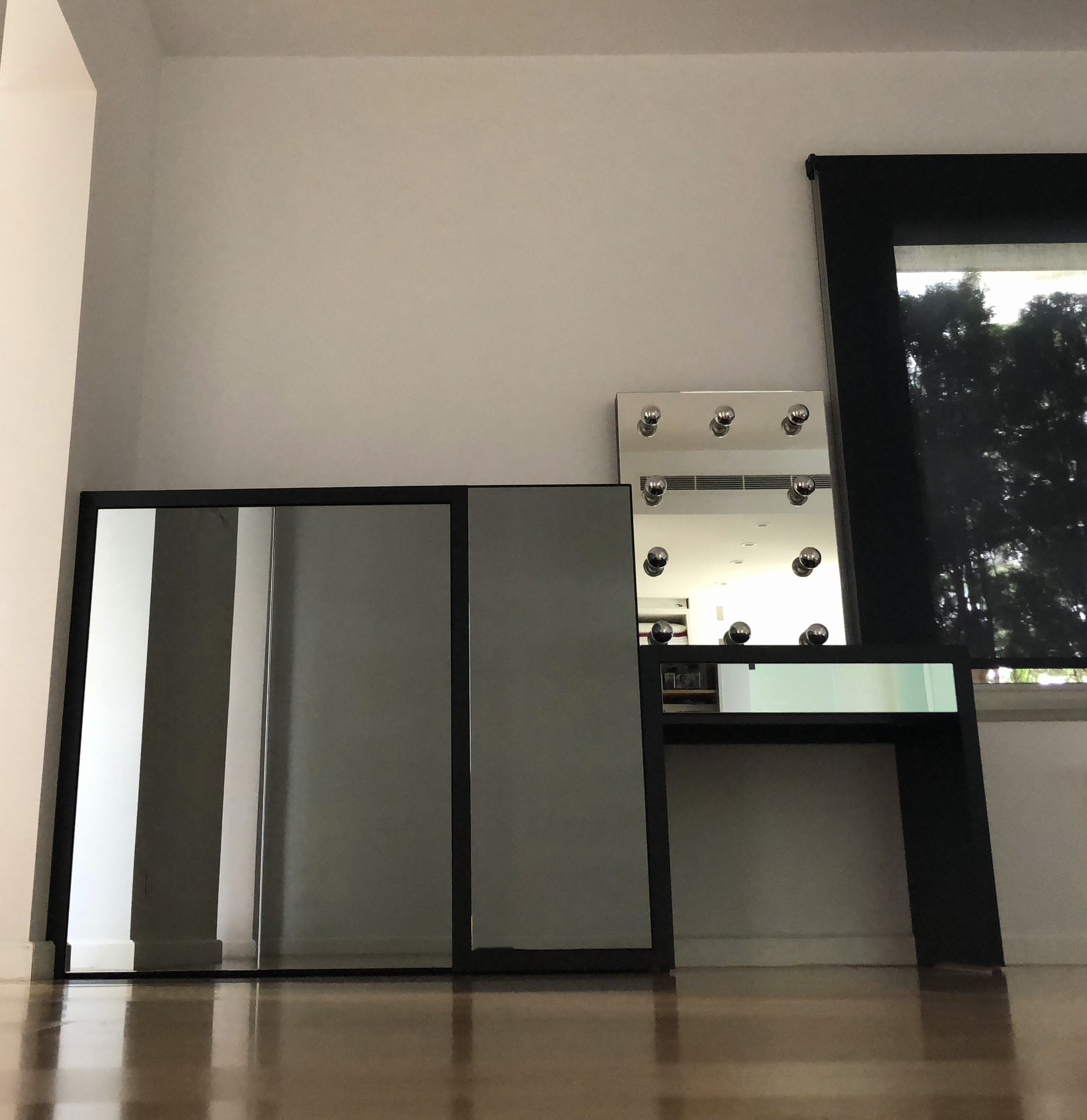 Interior of a room with a black-framed mirror, a set of vanity lights, a white mini heater, and a window with a view of trees outside.