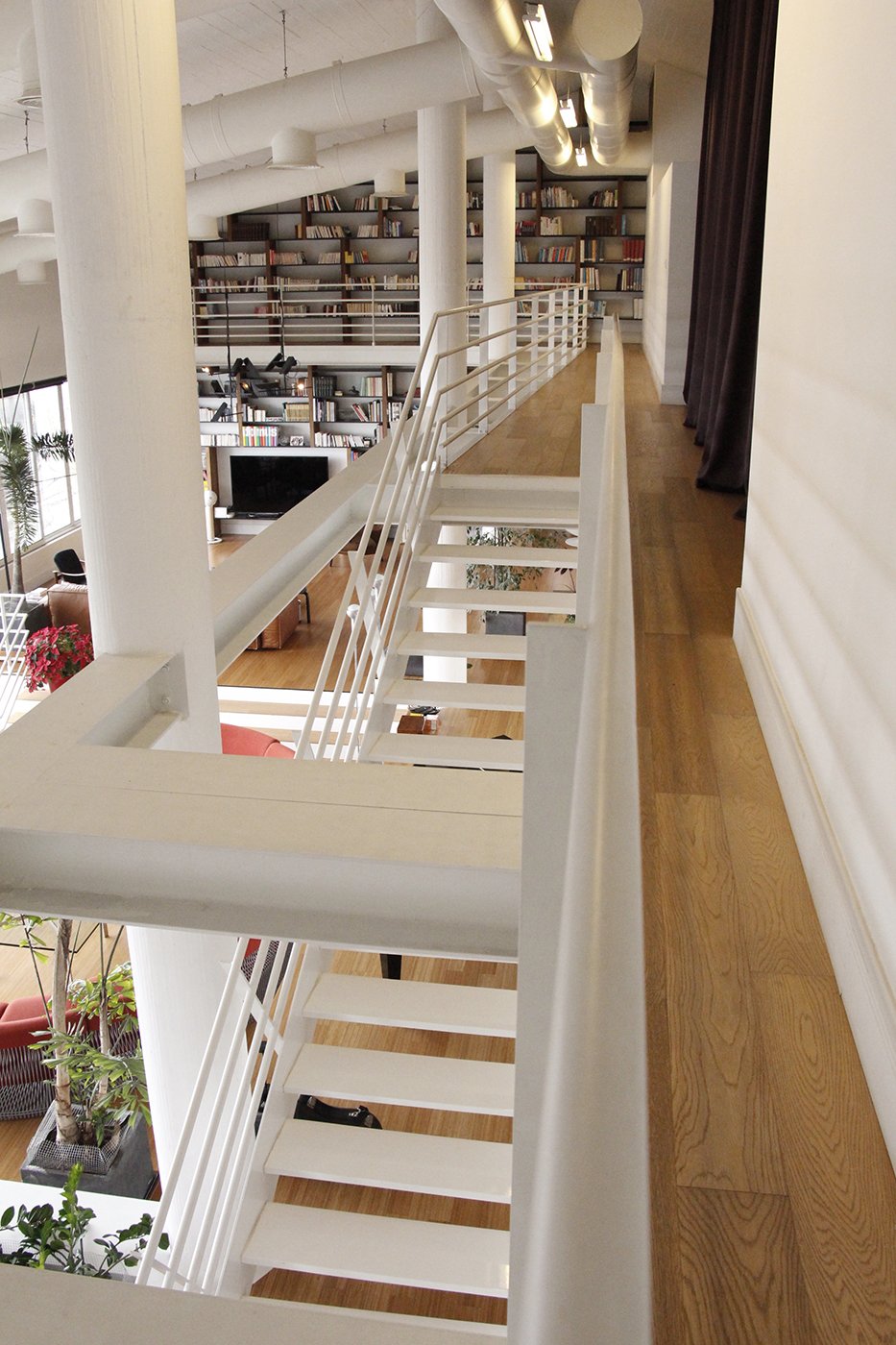 An indoor loft area with wooden flooring, white staircase, railing, and large bookshelves filled with books. The lower level has a seating area with plants, a couch, and a TV.