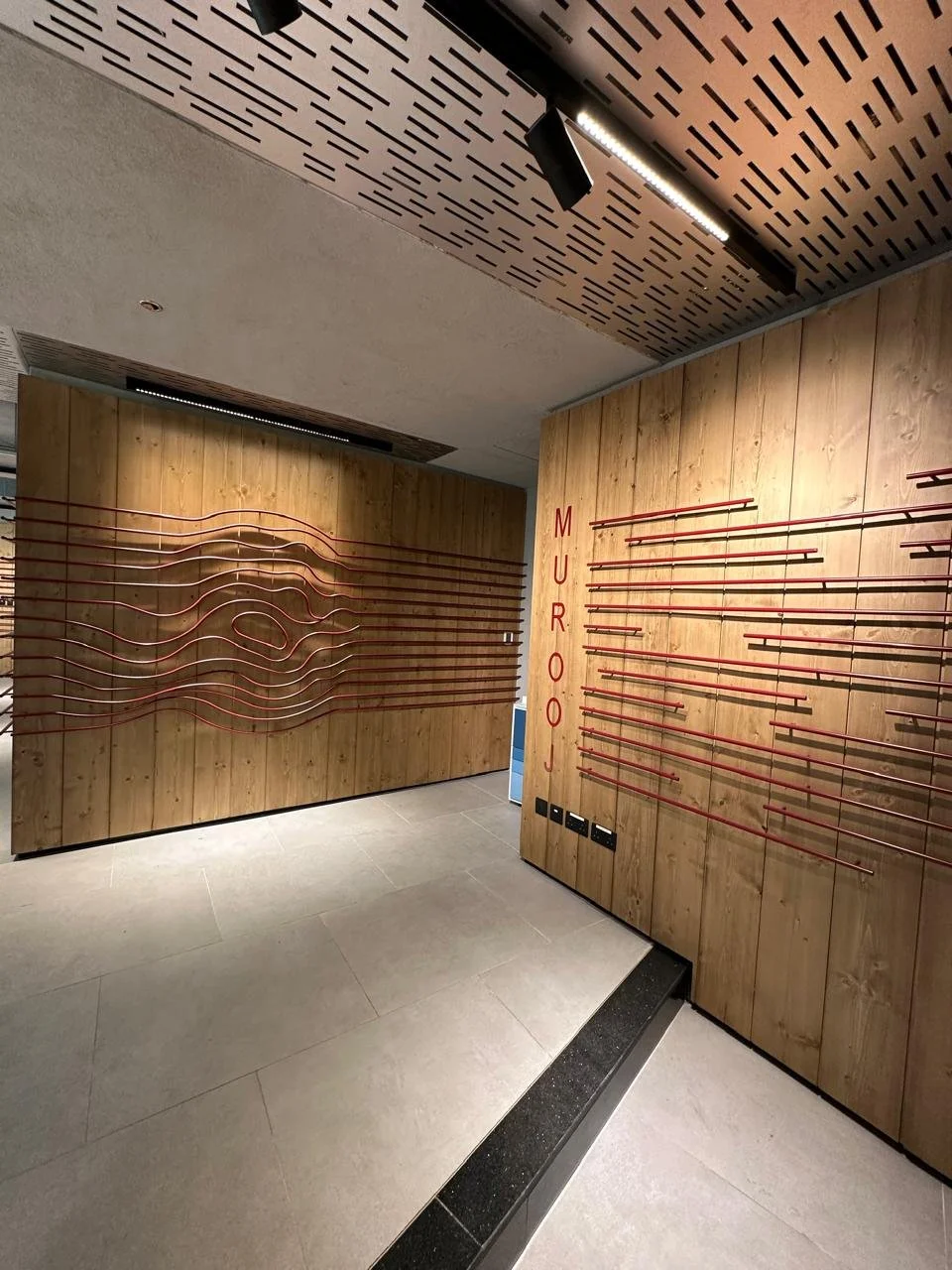 Interior of a modern store with wooden walls and red display racks. The left wall has a sculptural design of curved metal lines, and the right wall has horizontal red bars. The name "MUROU" is vertically displayed on the right wall in red lettering.