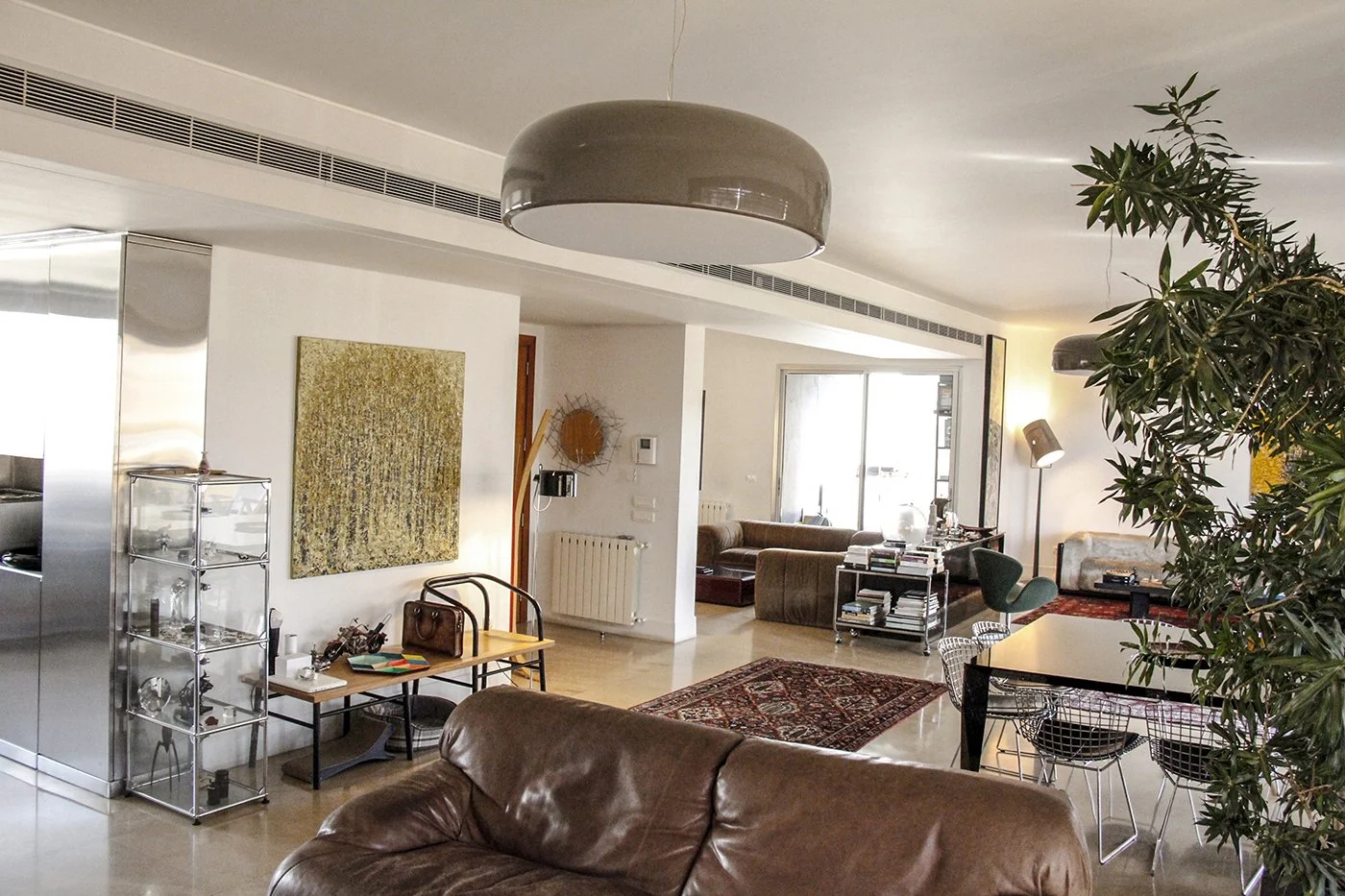 Spacious living room with brown leather sofa, dining table with wire chairs, bookshelves, and large windows.