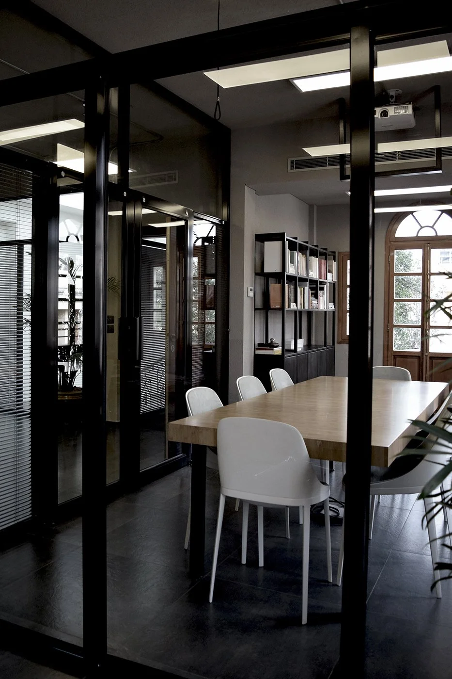 Modern office conference room with a large wooden table, white chairs, black metal framing, bookshelf, and natural light from windows.