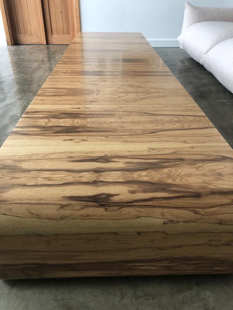 Long wooden table with a natural finish, located in a room with a concrete floor, a beige sofa, and wooden doors.