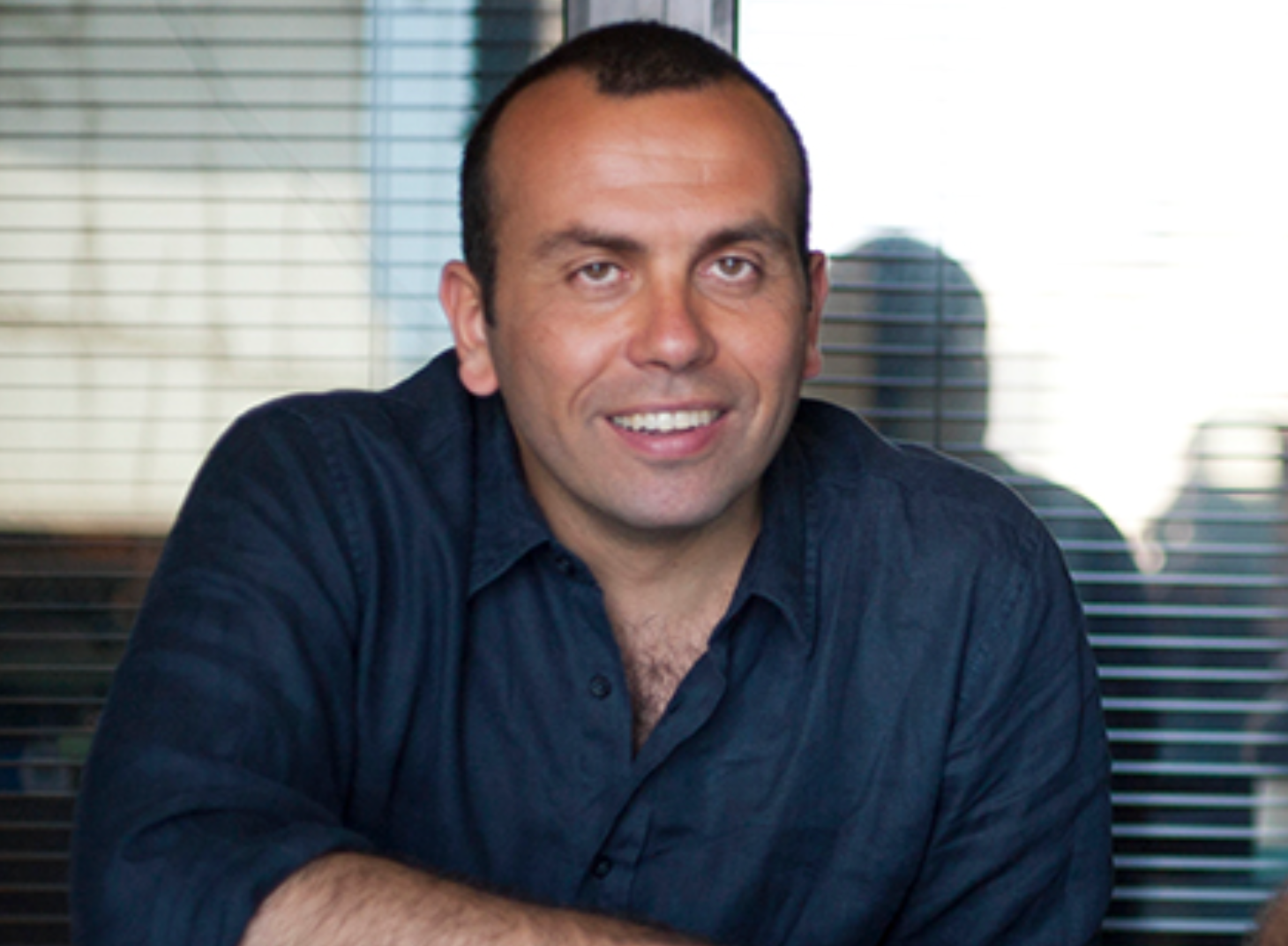 Man smiling while sitting at a table in an office setting with blinds in the background.