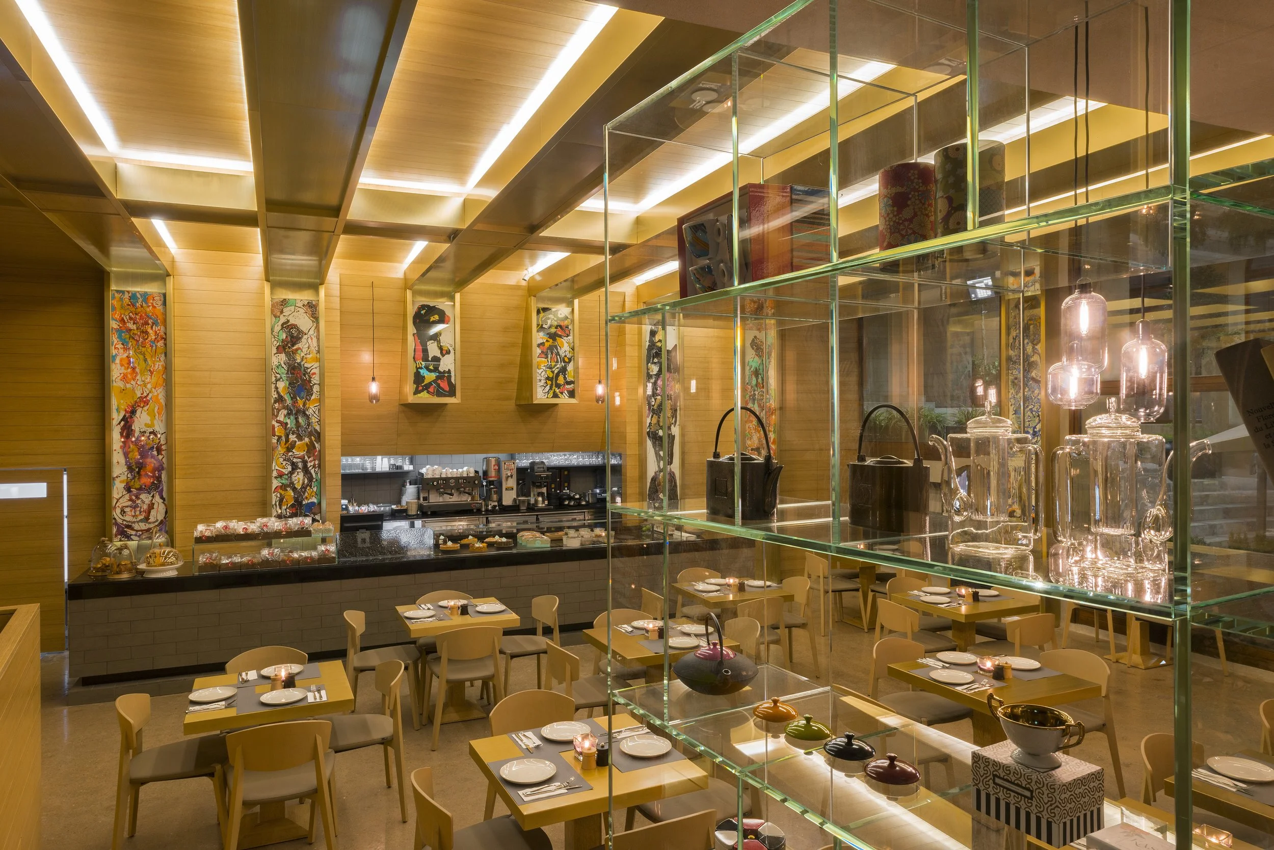 A restaurant interior with wooden walls and ceiling, decorated with colorful abstract paintings. There are multiple tables set with plates and napkins, and a glass display case with glassware and ceramic vases. Pendant lights hang from the ceiling, a