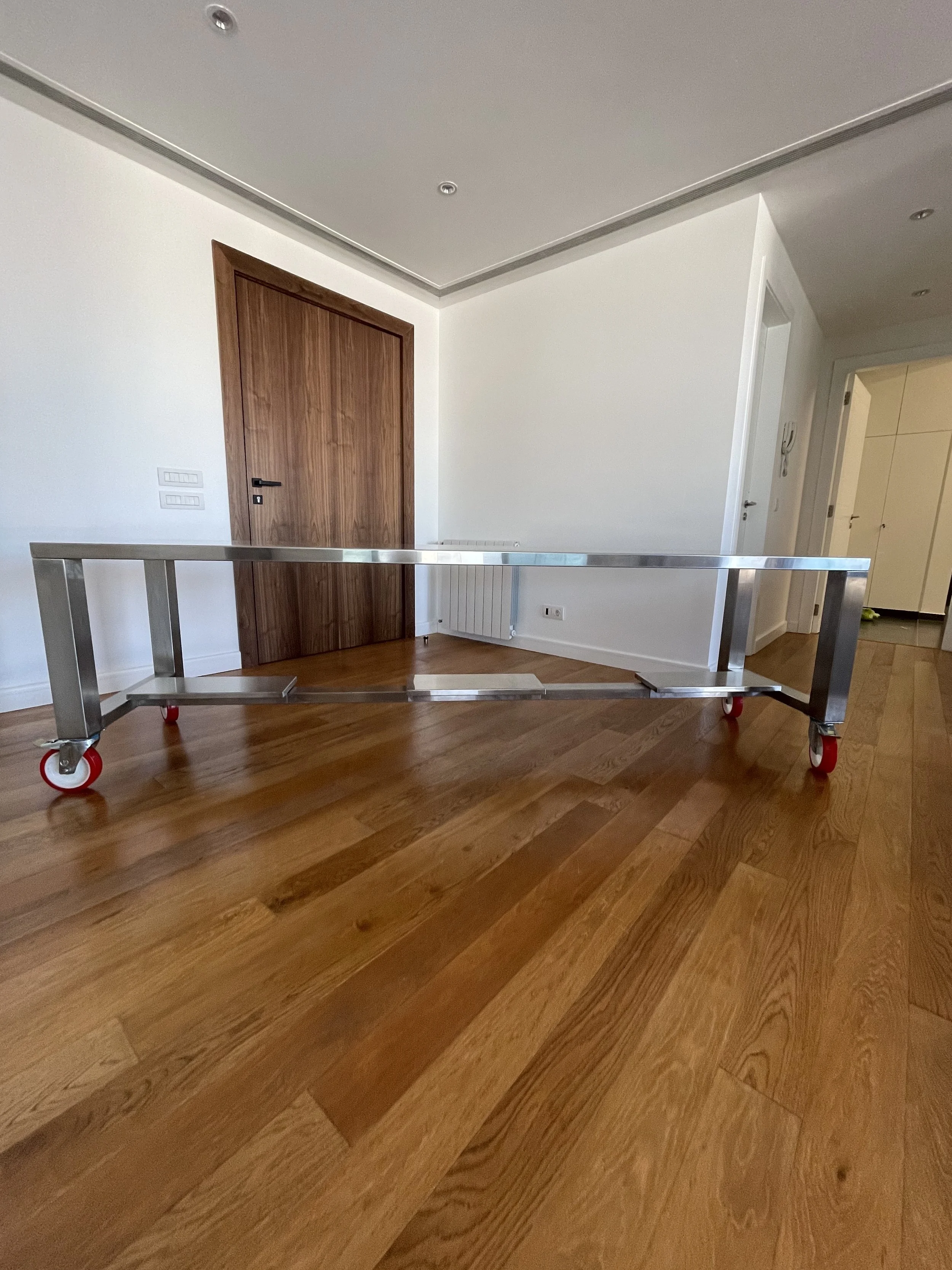 Empty room with white walls, a wooden door, wooden flooring, a metal handrail on casters, and an open doorway leading to another room.