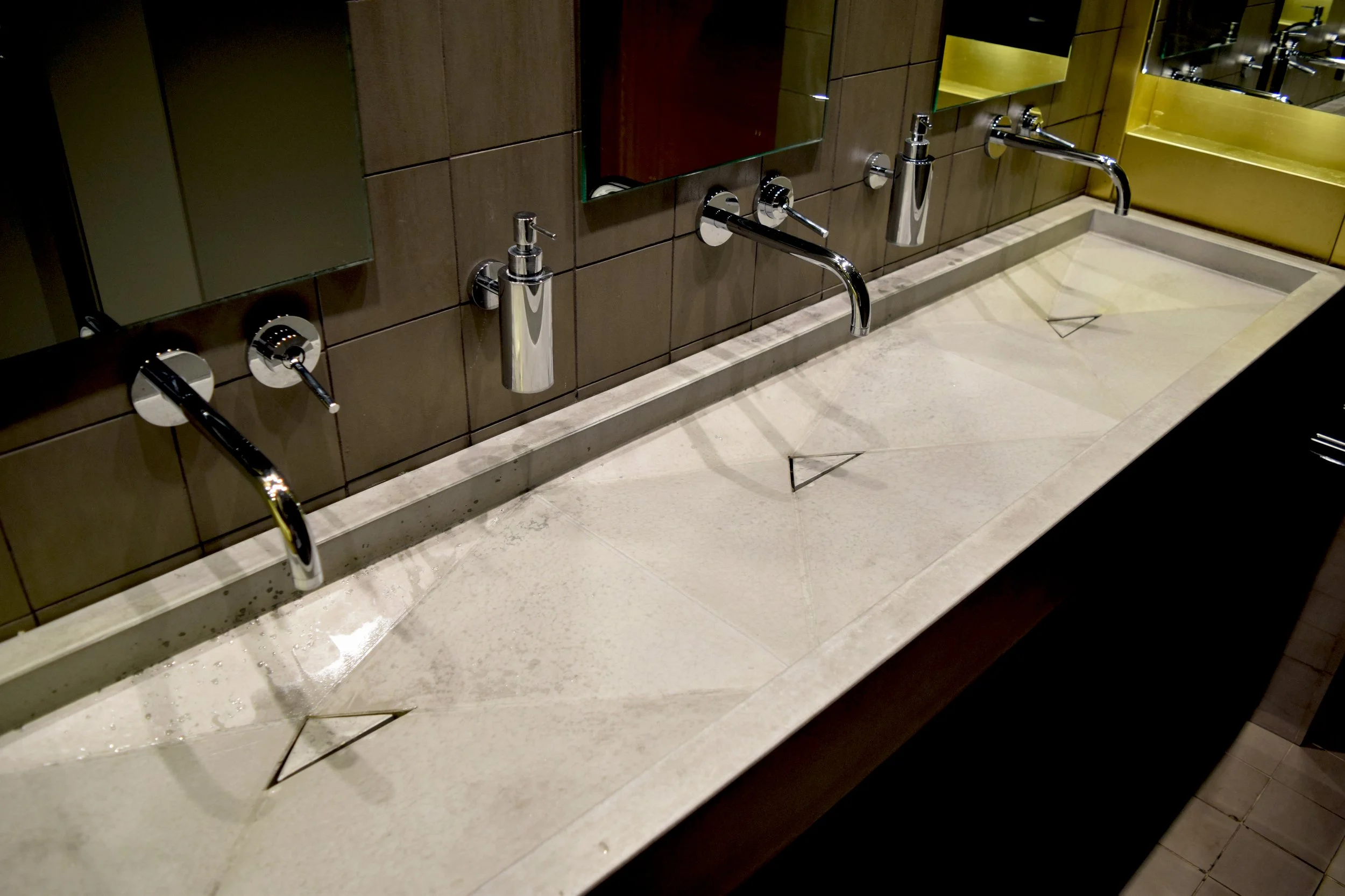 Restroom sinks with chrome fixtures, brown tiled wall with mirror, and beige countertop in a public bathroom.