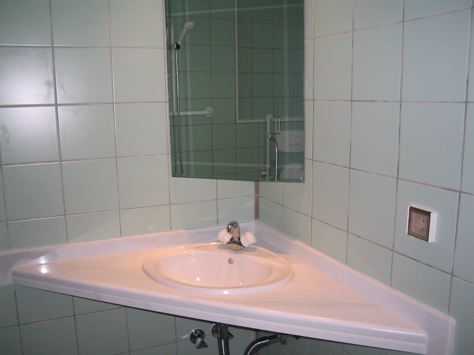 Bathroom sink with beige marble top, faucet, and two white handles, with mirror and green tiled shower area in background.