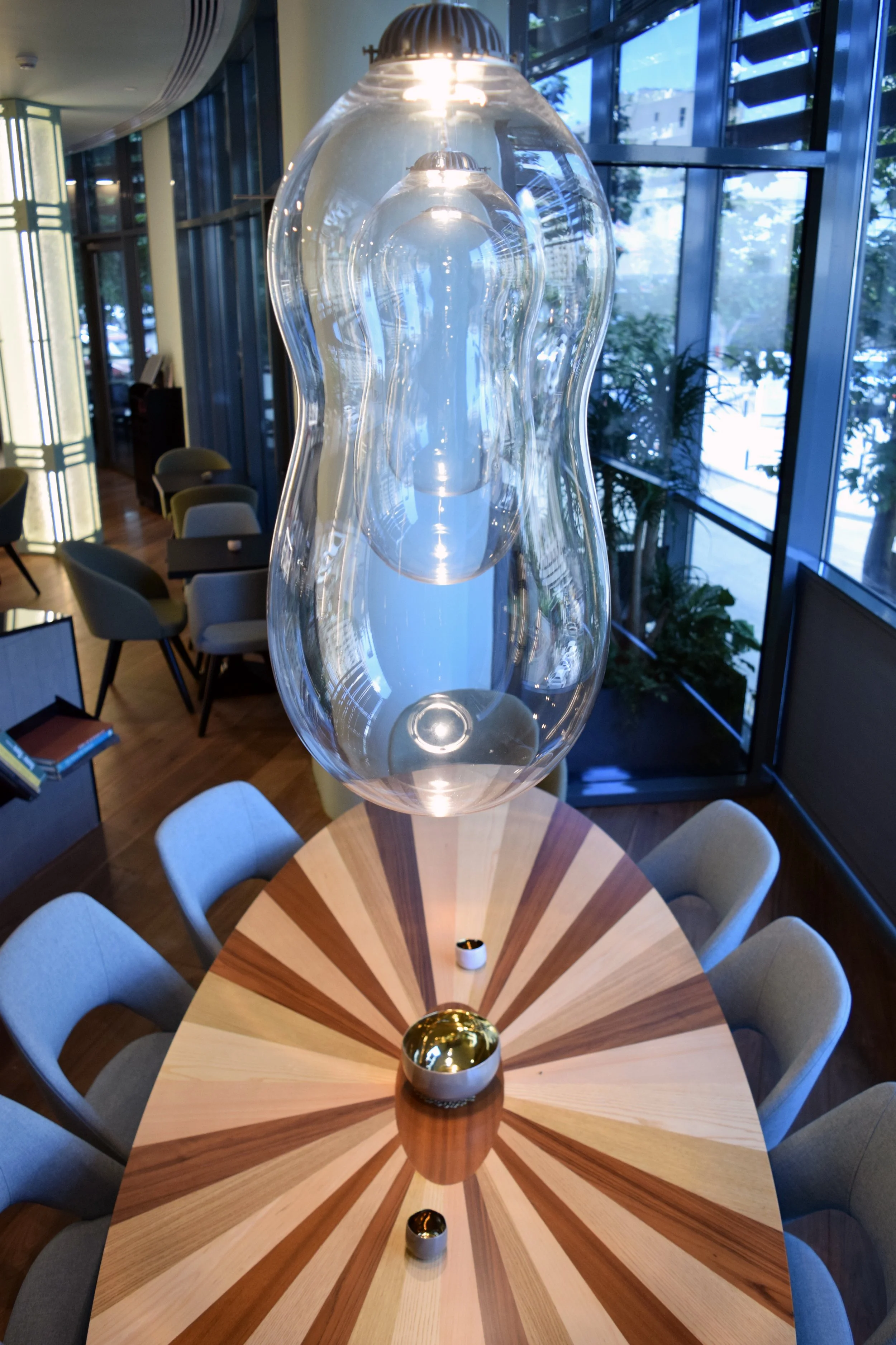 Close-up view of a modern hanging glass light fixture in a stylish room with large windows, a round wooden table with a colorful radial pattern, and chairs around it.